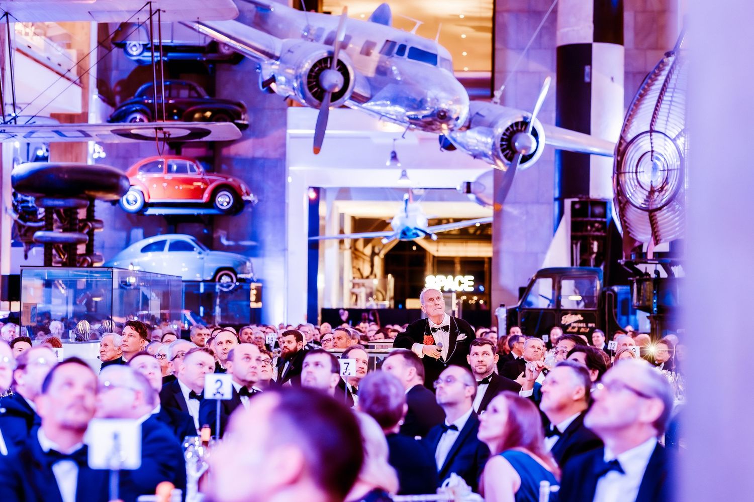 The science Museum Elegant gala event with guests in formal attire beneath vintage cars and aircraft displays in a grand venue.