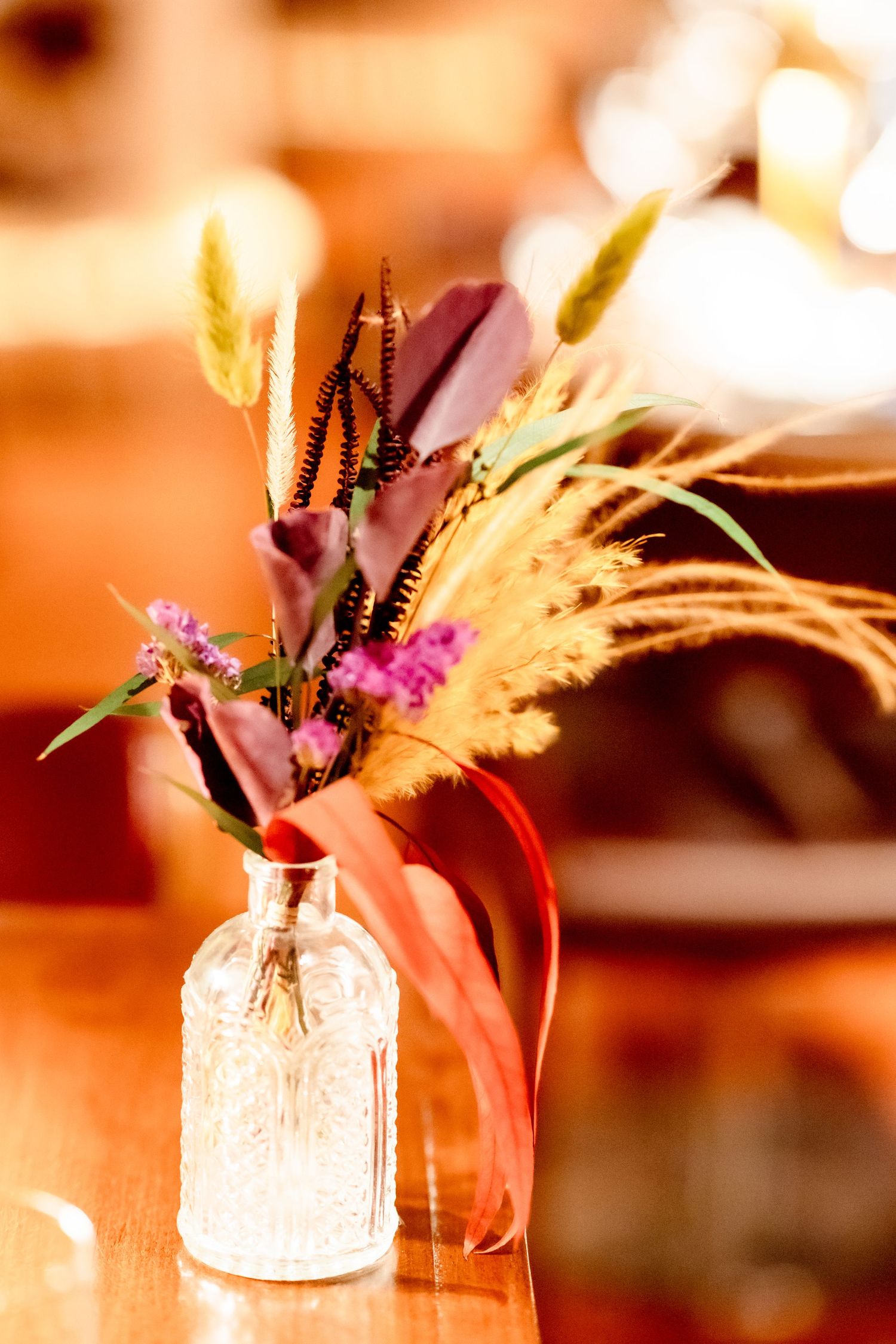 Small glass bottle vase with autumn dried flowers, wheat stems, and burgundy ribbon on a wooden table.