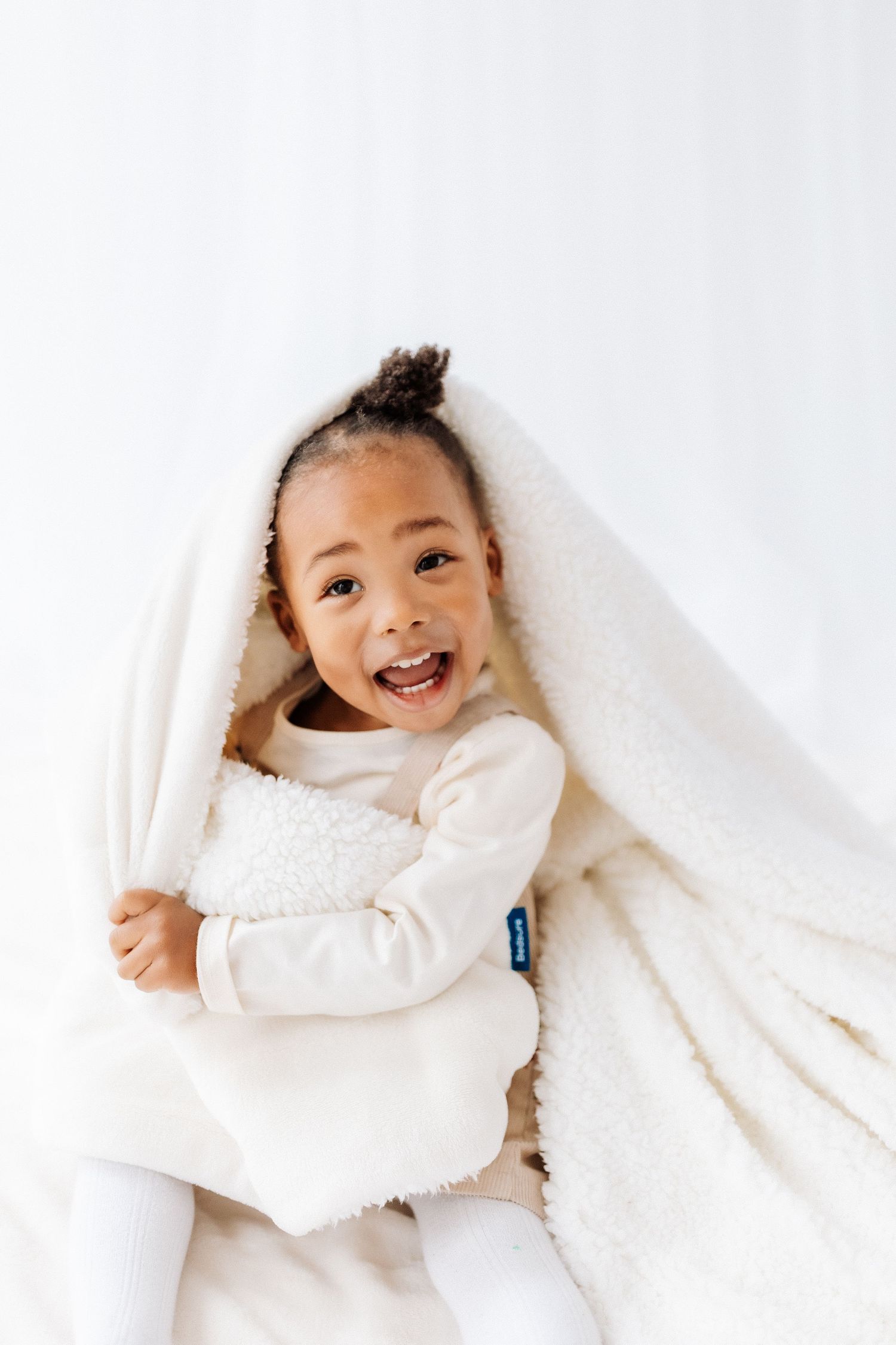 A happy child wrapped in a cozy white blanket smiles brightly against a light background. Shepperton Surrey photography studio portrait baby