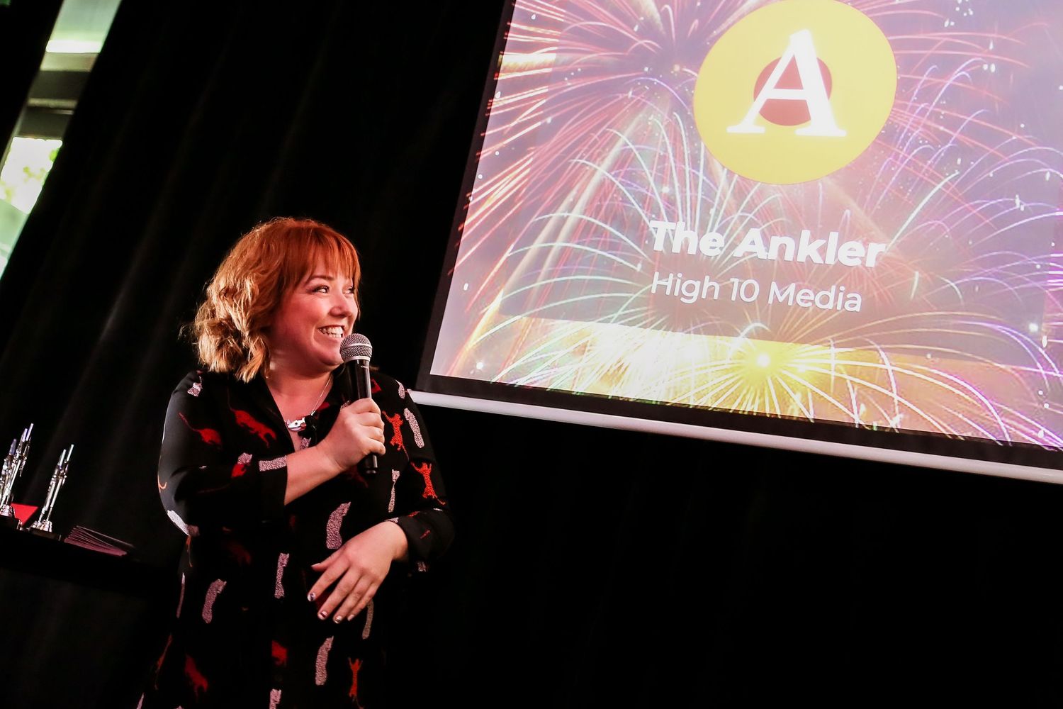 Woman speaking at at podcast awards in London holding microphone on stage with colorful fireworks display projected behind her.