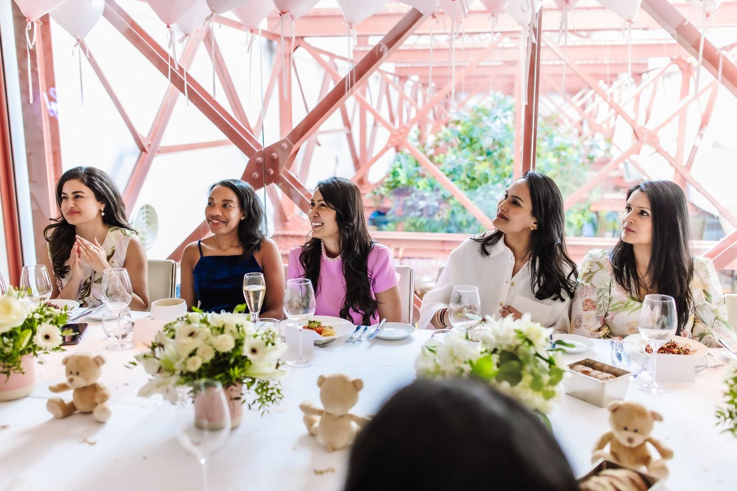 Five women laughing and celebrating at an elegant baby shower luncheon with floral centerpieces and teddy bear decorations.