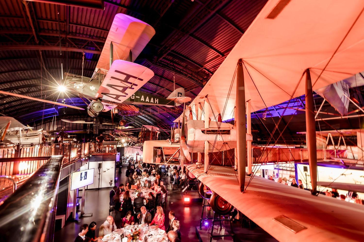 Elegant gala event in a museum hall with vintage aircraft displays, glowing pink lights, and guests dining at round tables.