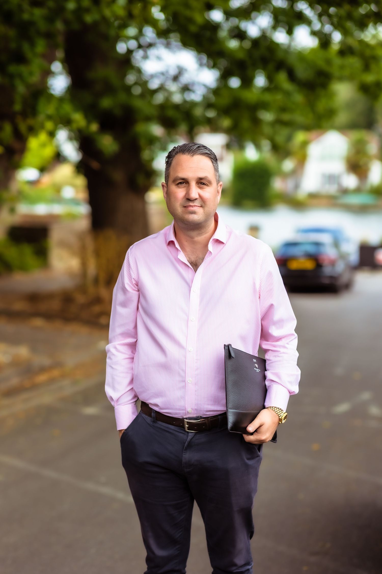 Professional in a pink dress shirt and dark trousers walking confidently down a tree-lined street.