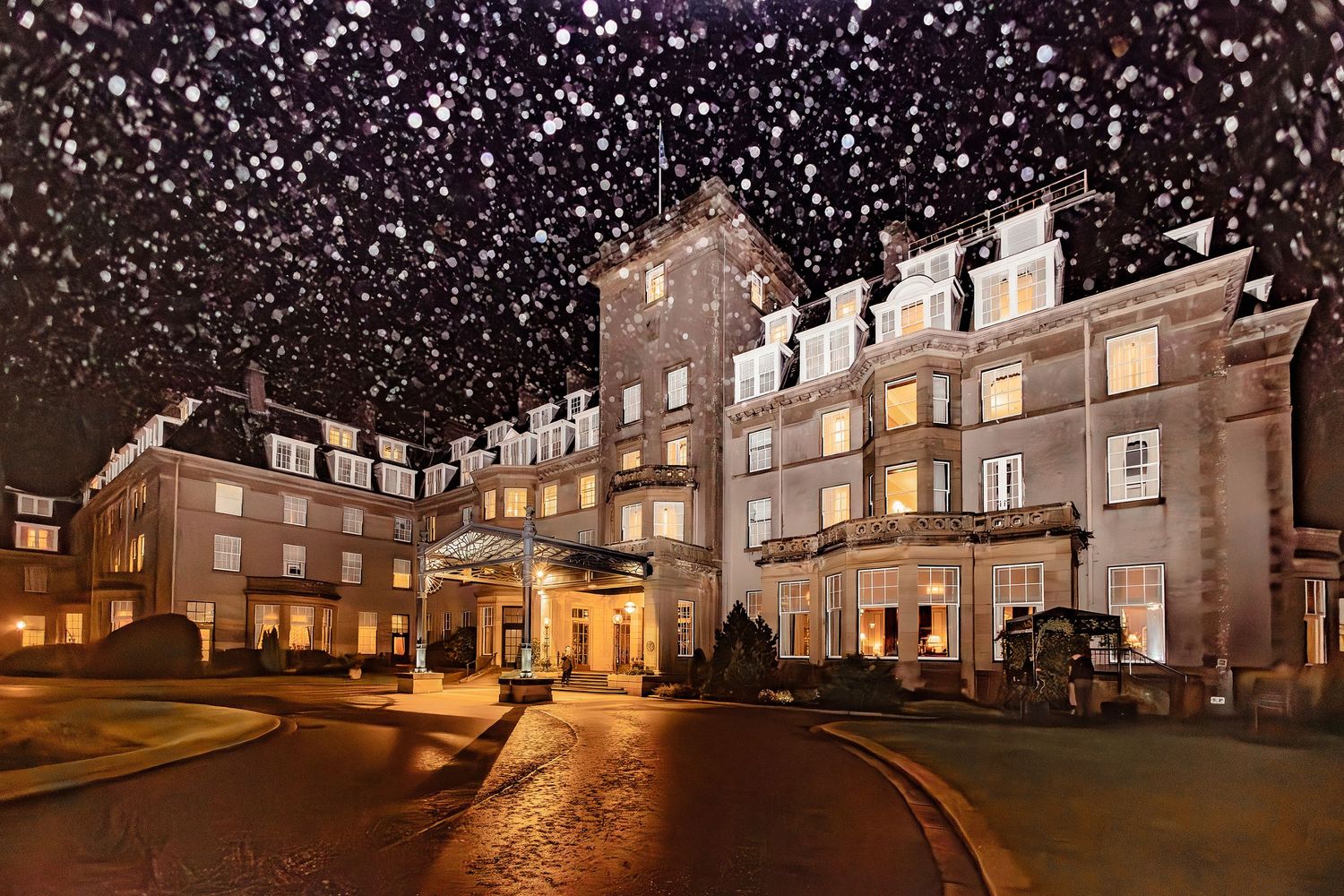 Elegant hotel exterior illuminated at night with snow falling, warm golden lights glowing from windows in winter scene.