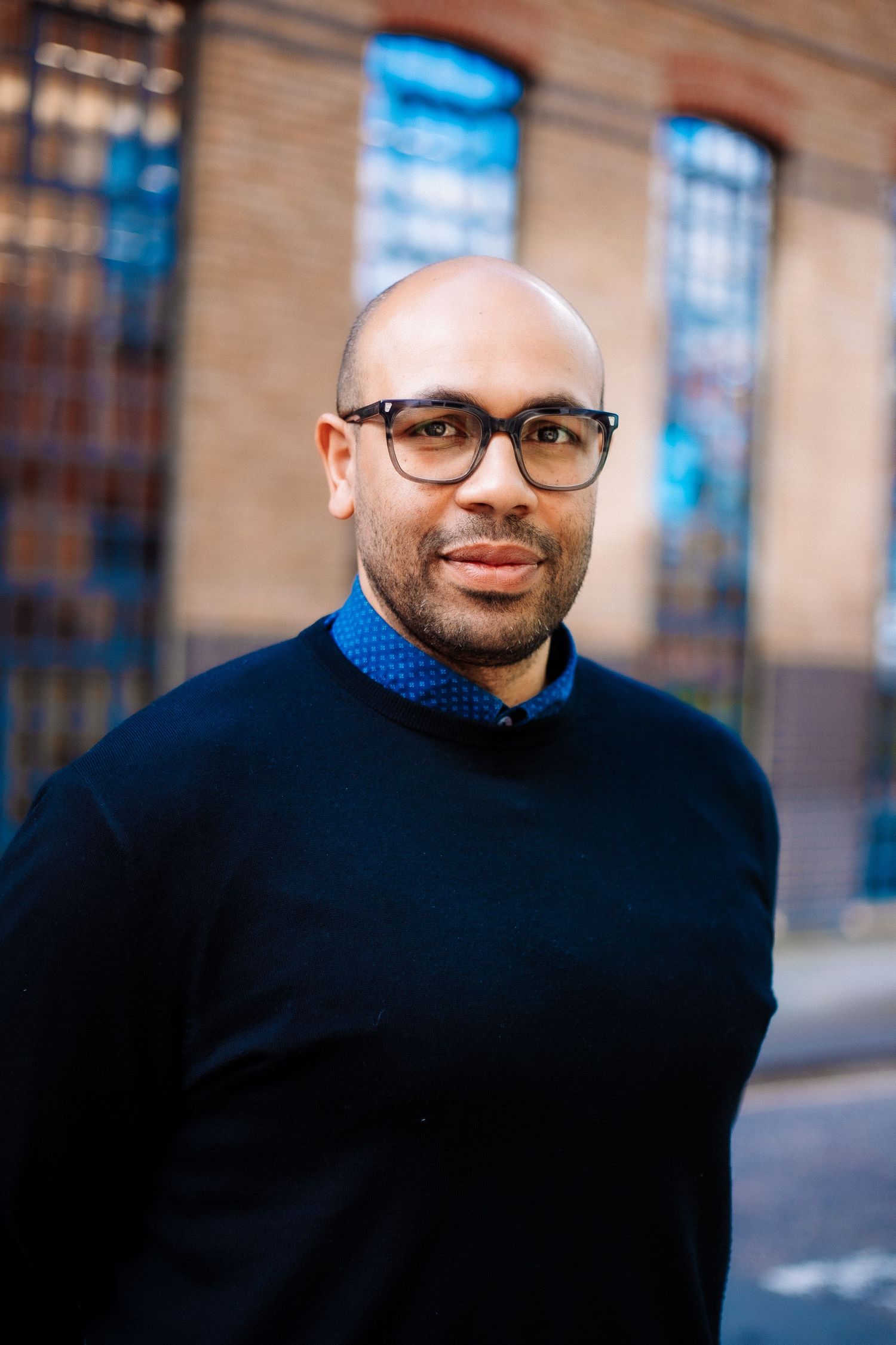Professional headshot of someone wearing glasses and a navy sweater standing outdoors against a brick building.