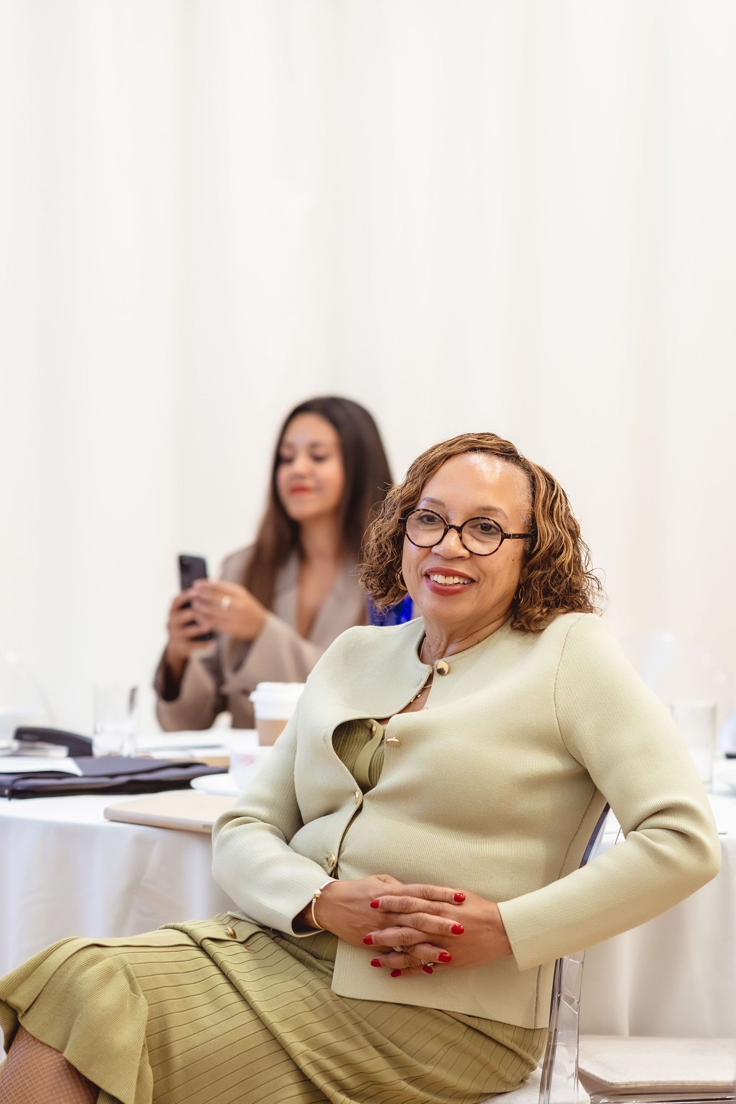 Professional woman in glasses and blazer smiling at camera during business meeting, colleague using phone in background.