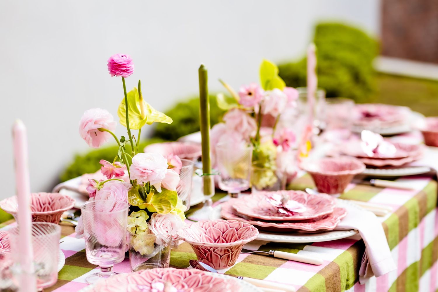 Elegant pink and green table setting with floral centerpiece, pink plates, and candles on a checkered tablecloth.