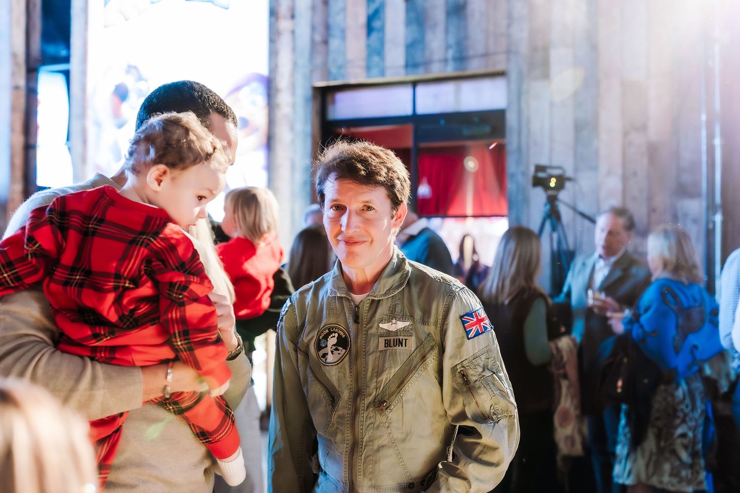 Military pilot in olive flight suit holds child in red plaid shirt at indoor evening event.