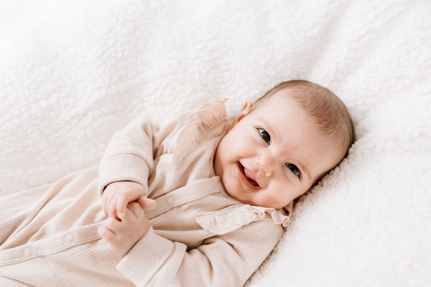 A baby in white clothing lies on soft white bedding while looking to the side.