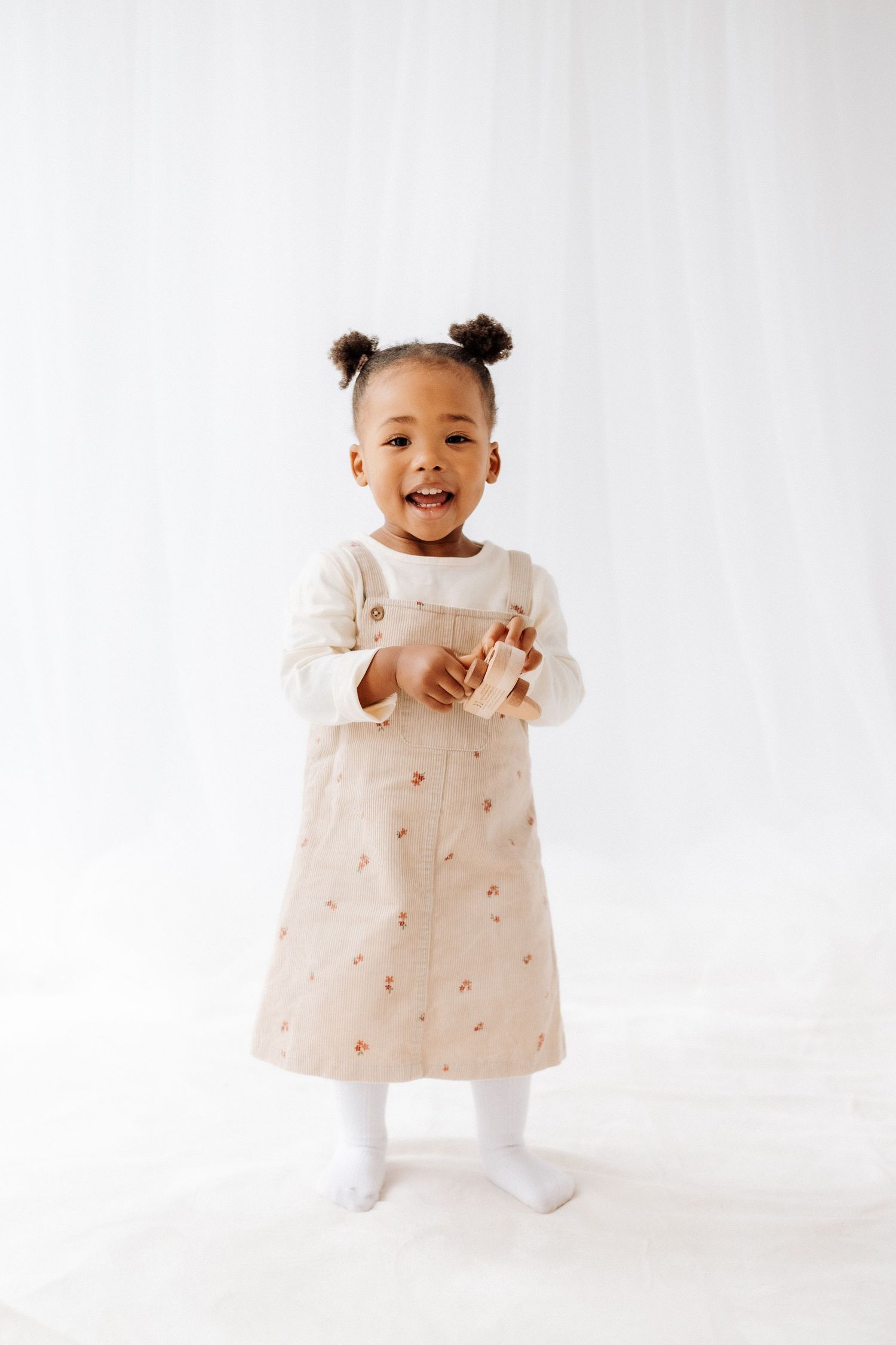 A baby in a dress with floral details stands against a white background in a three-photo sequence. Shepperton Surrey photography studio