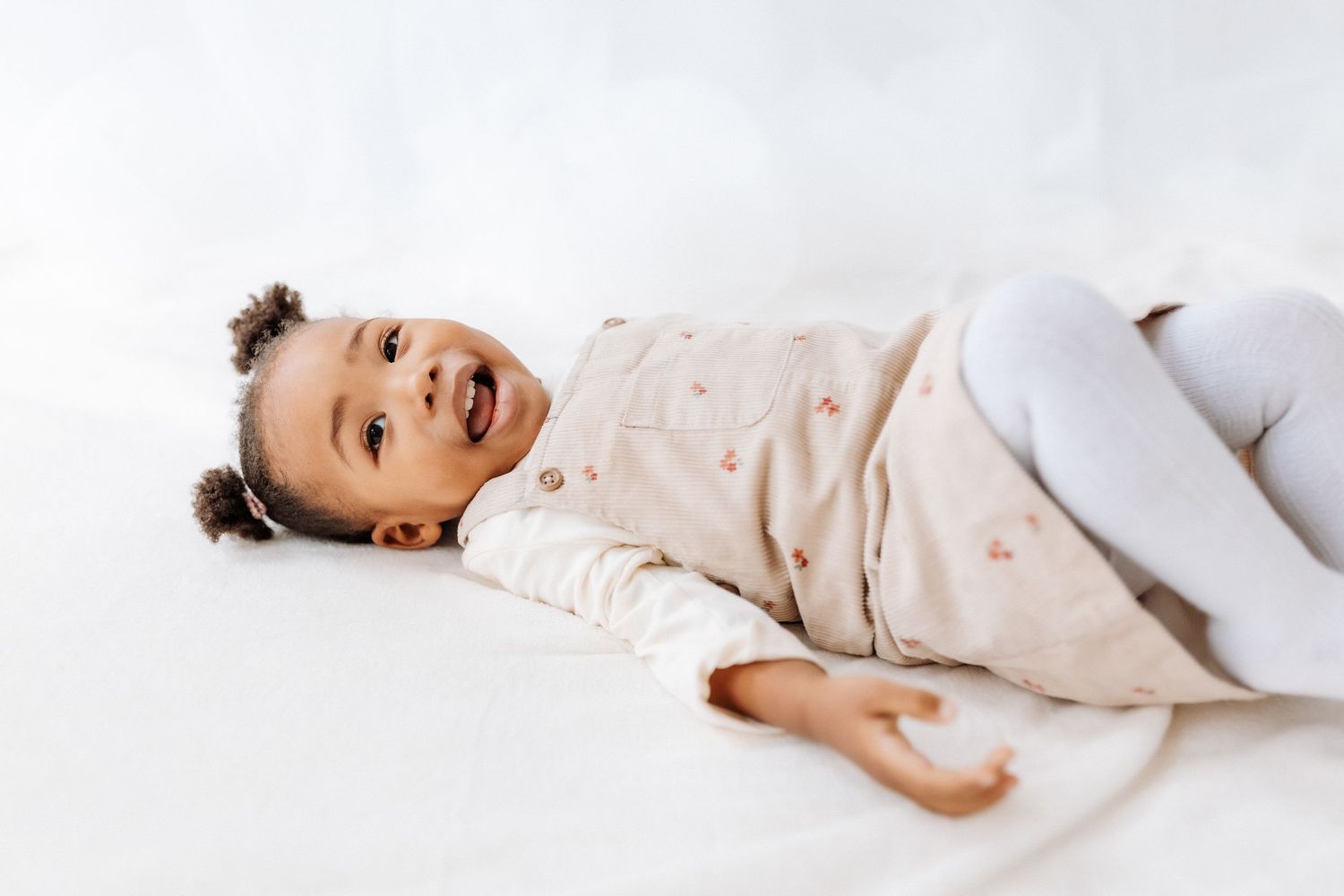 A joyful child wearing a light colored outfit lies on a white surface with a playful expression.