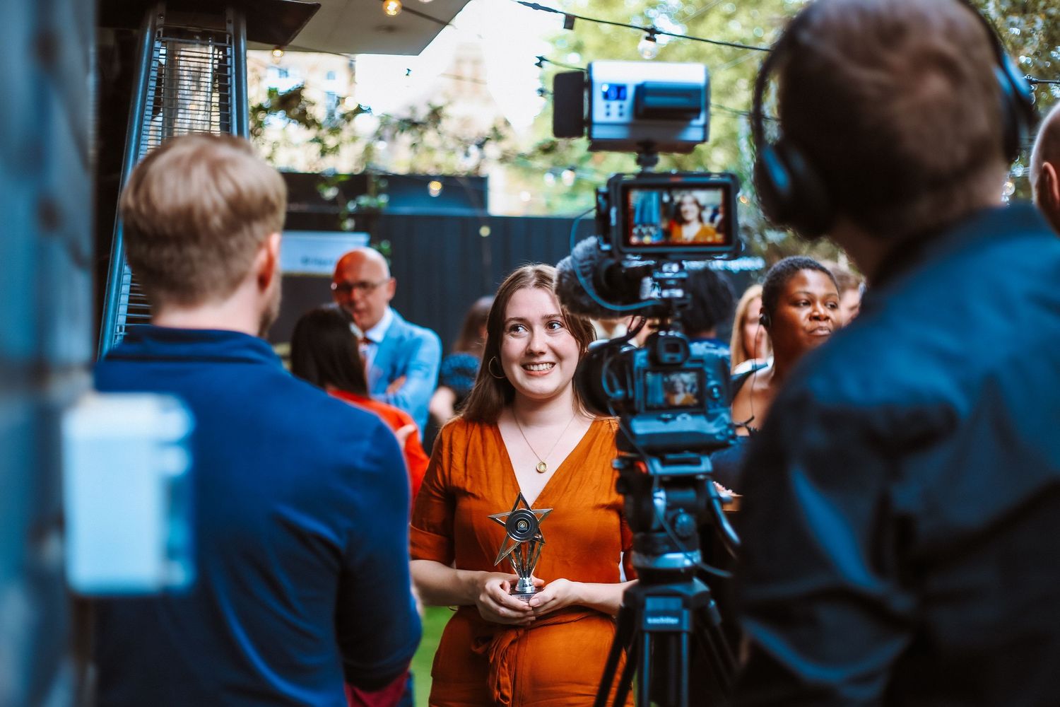 Woman in orange dress being filmed by a professional camera crew at an outdoor evening event. award ceremony interview photography