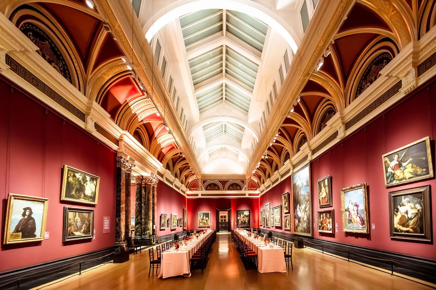 Elegant museum gallery with red walls, arched ceilings, skylight, and paintings displayed alongside white-clothed dining tables.