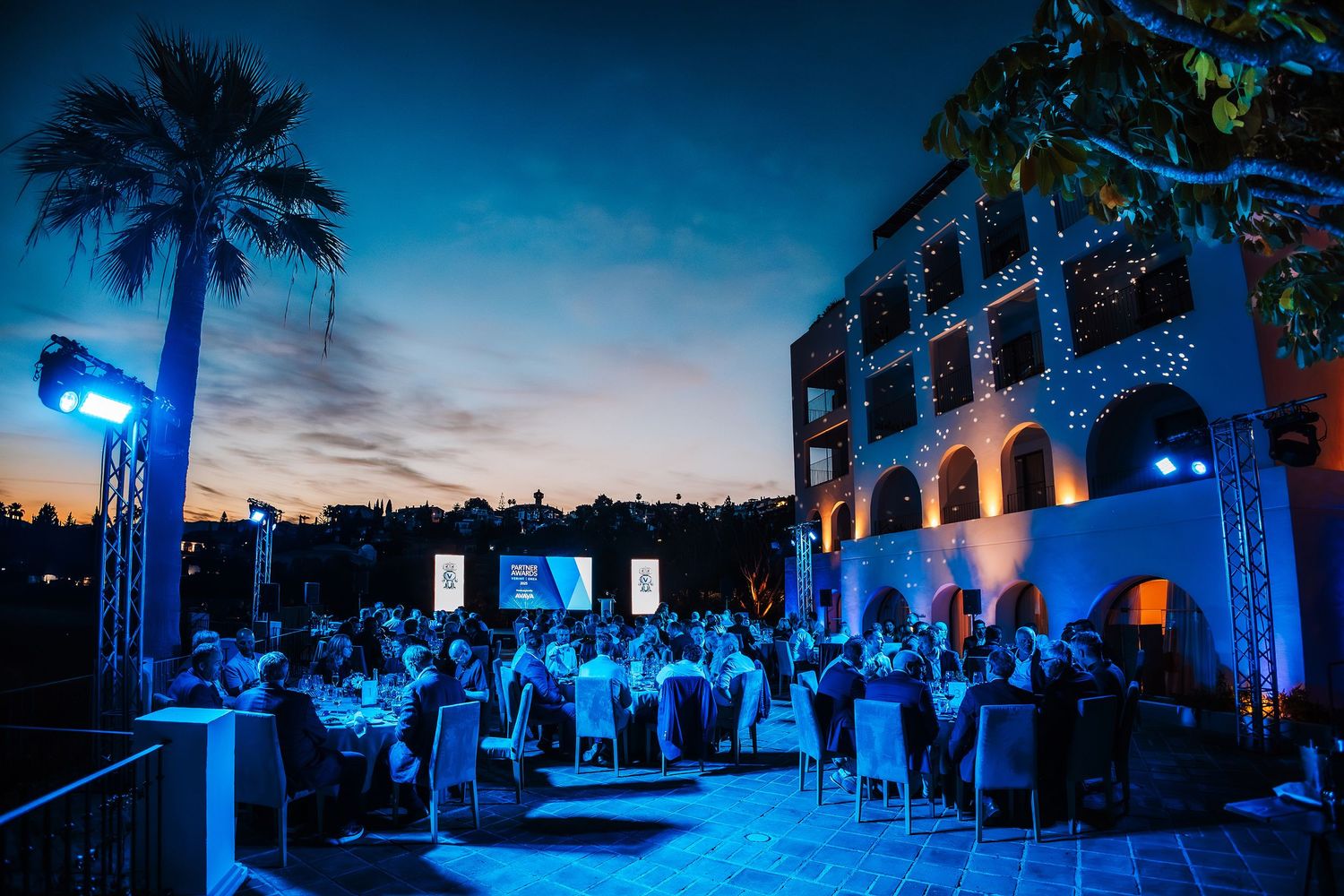 Elegant outdoor evening gala dinner at a white Mediterranean villa illuminated in blue light at dusk.