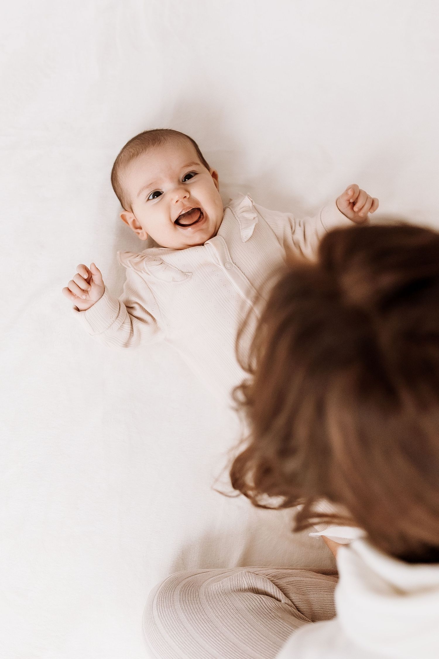 Baby lying on white surface smiling and laughing with arms raised while being watched from above. candid photography baby, family Shepperton