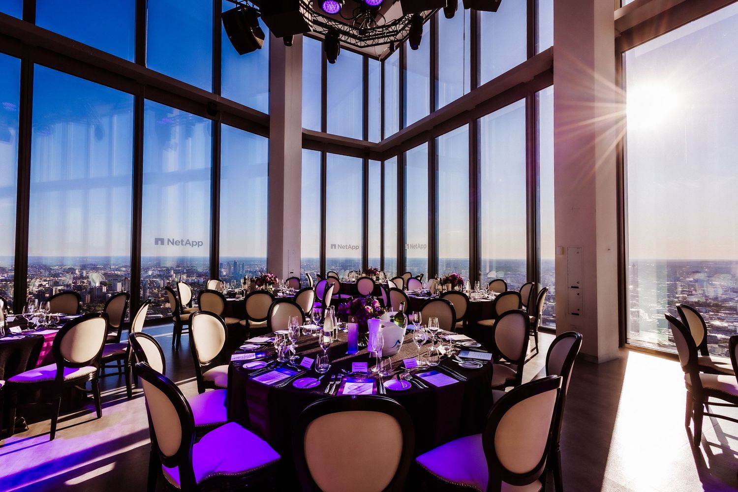 Elegant rooftop banquet hall with purple lighting, round tables, and panoramic city views through floor-to-ceiling windows.