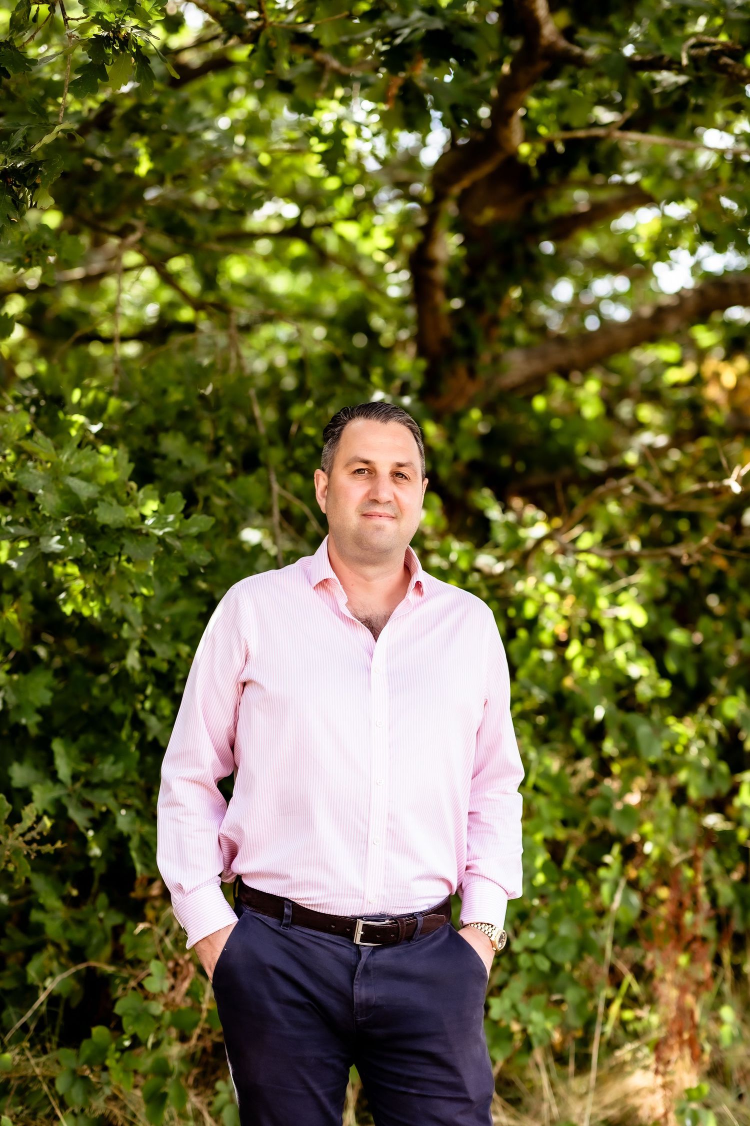 Person in pink dress shirt and navy pants stands casually against lush green foliage background in outdoor setting.