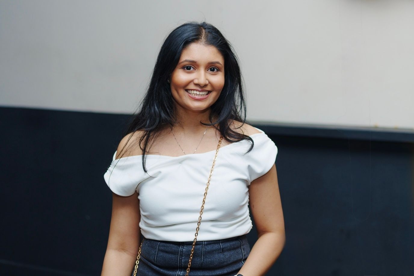 Young woman in white off-shoulder top smiles casually against a wall with a chain strap purse.