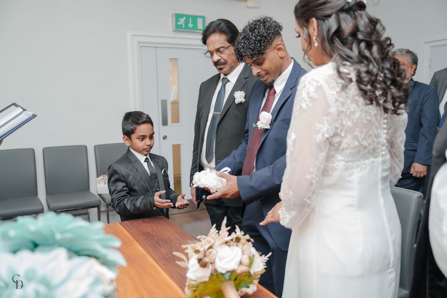 Wedding party gathered around table during document signing ceremony with decorative elements.