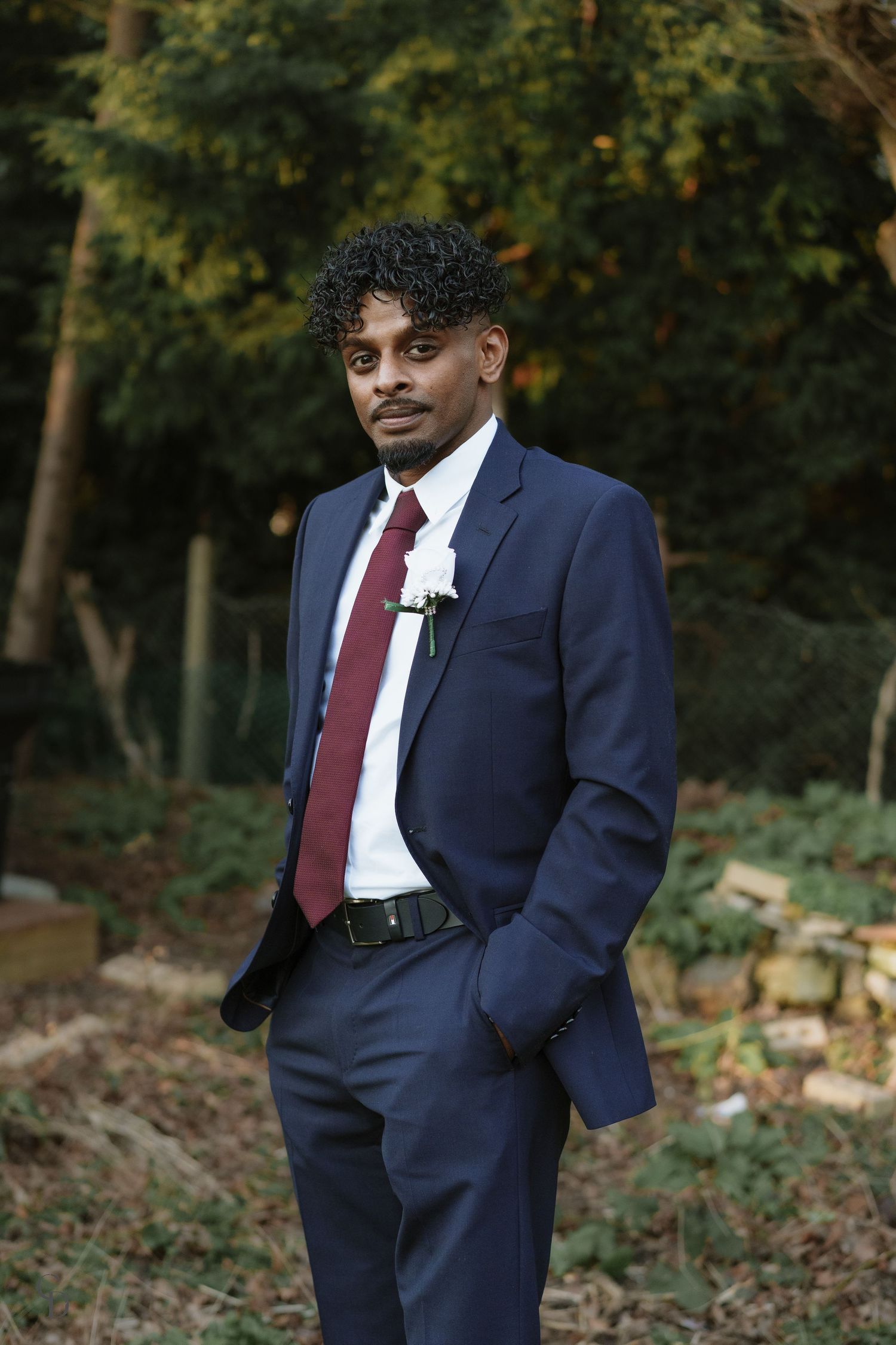Groom in navy suit and burgundy tie poses in outdoor garden setting.