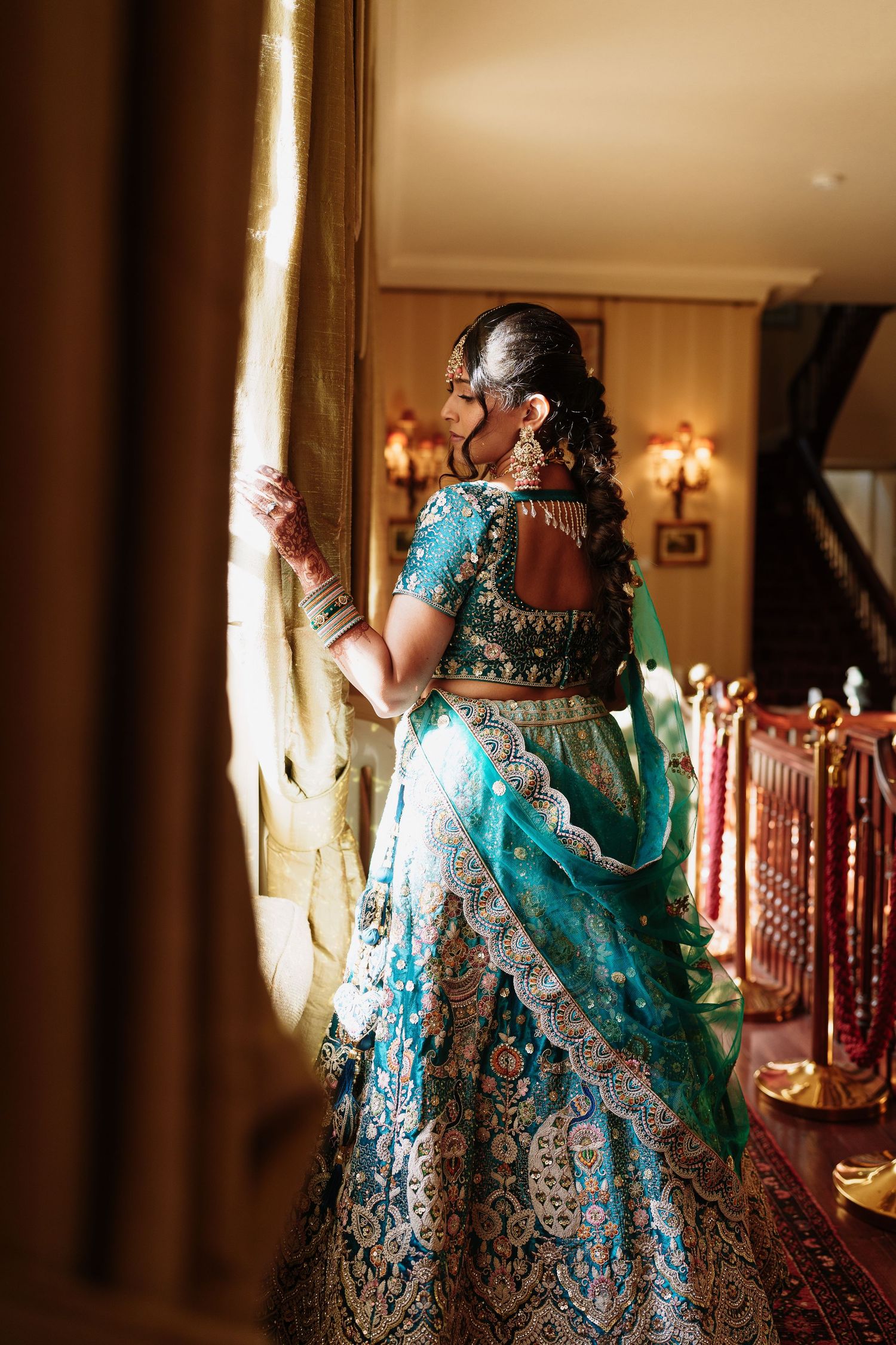 Indian bride in a stunning turquoise and gold embroidered lehenga stands near window with warm light streaming in.