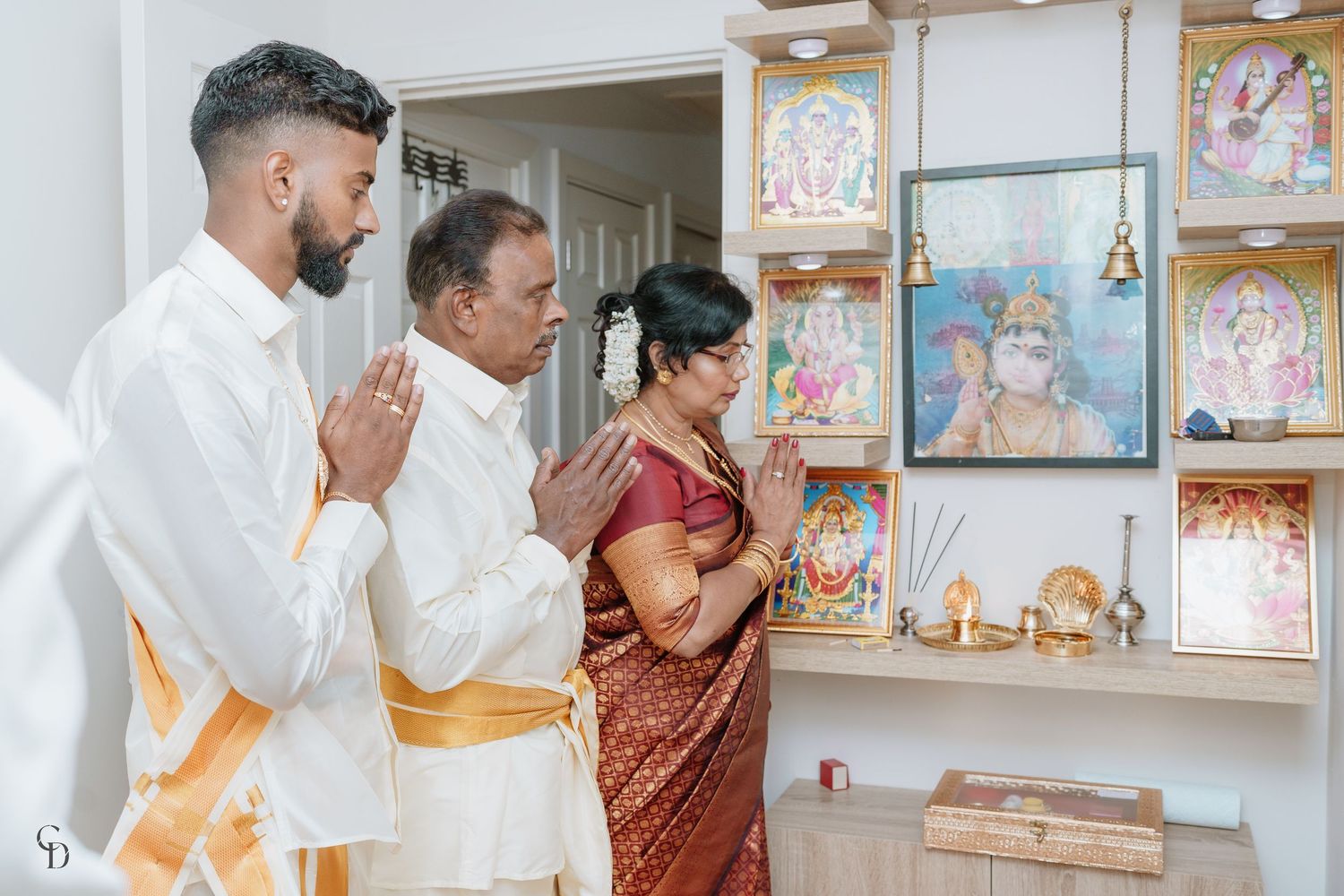 Three people praying at home shrine with religious artwork and decorations.