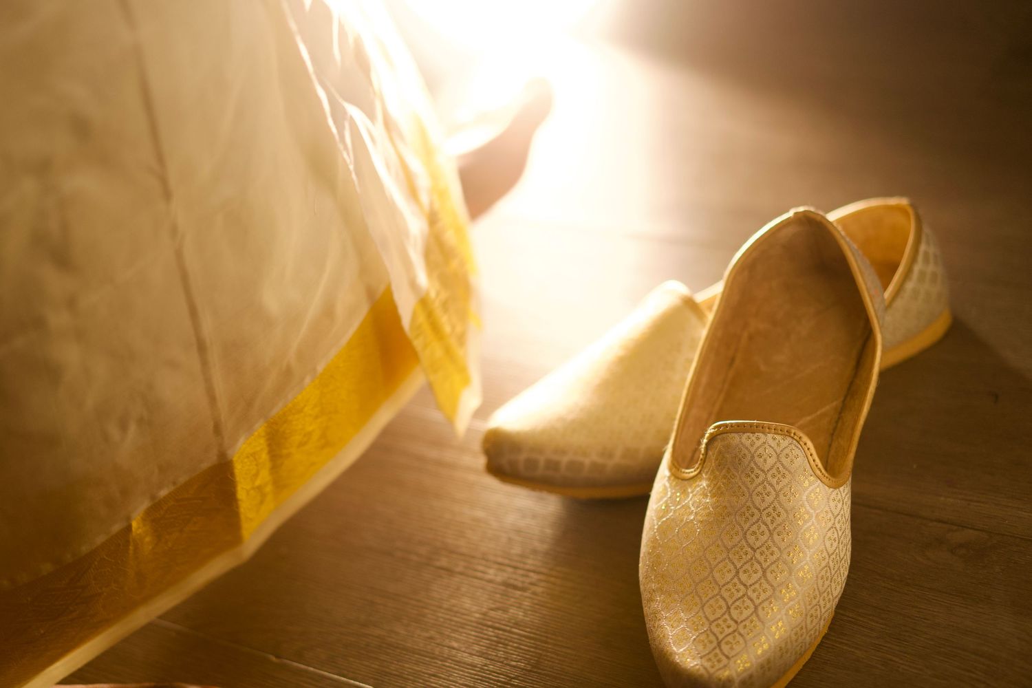 Golden espadrille slip-on shoes resting on wooden floor beside a white and yellow traditional outfit hem.