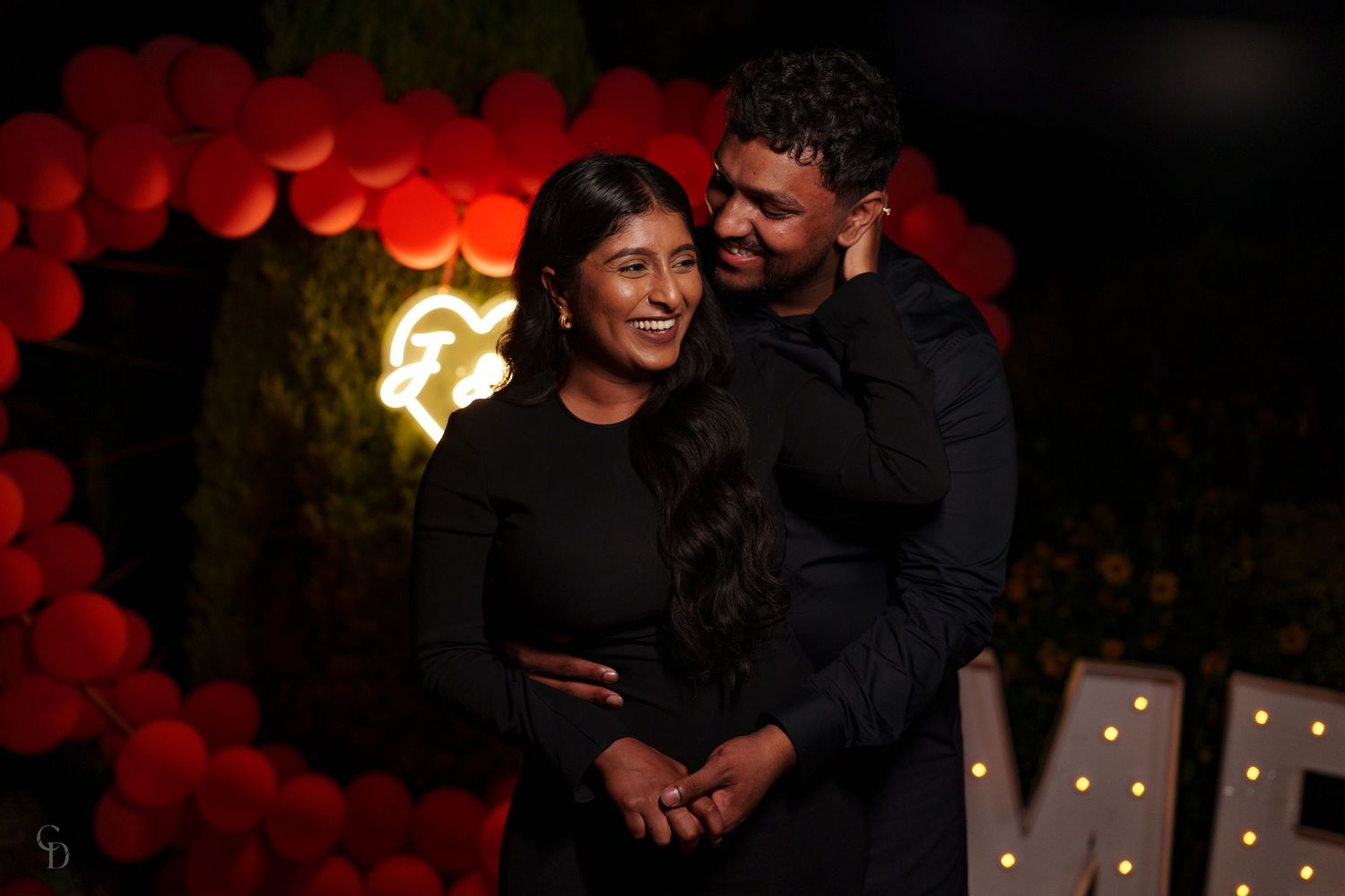 A romantic portrait with red balloon decor and soft lighting showing a couple in matching black outfits.