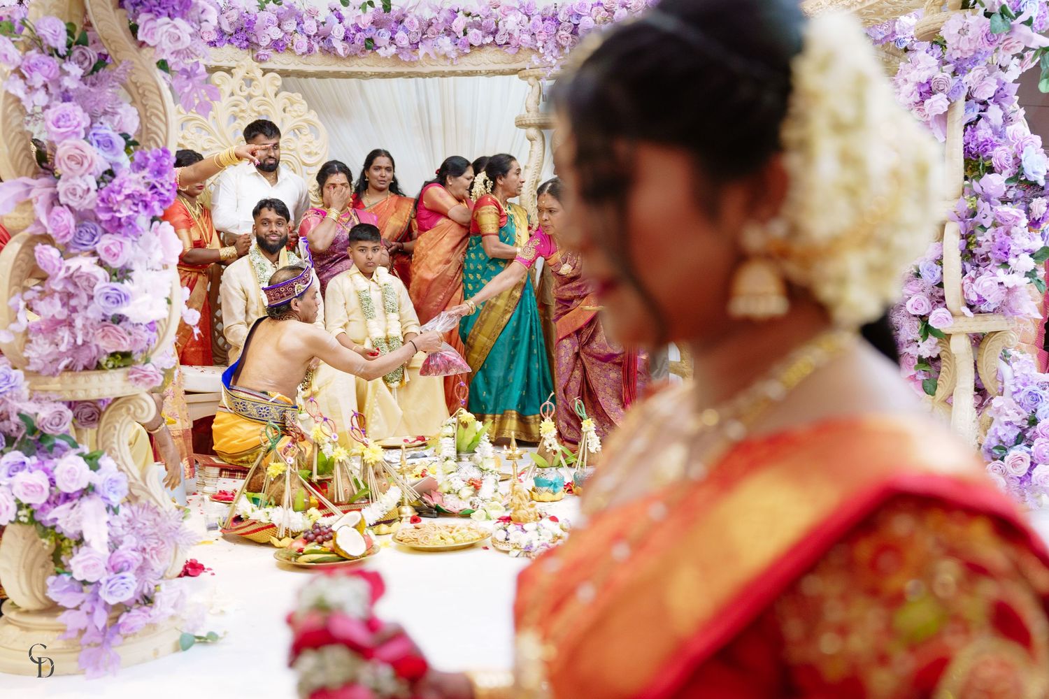 Traditional Indian wedding ceremony with guests gathered around decorated mandap with purple floral decor and ritual items.