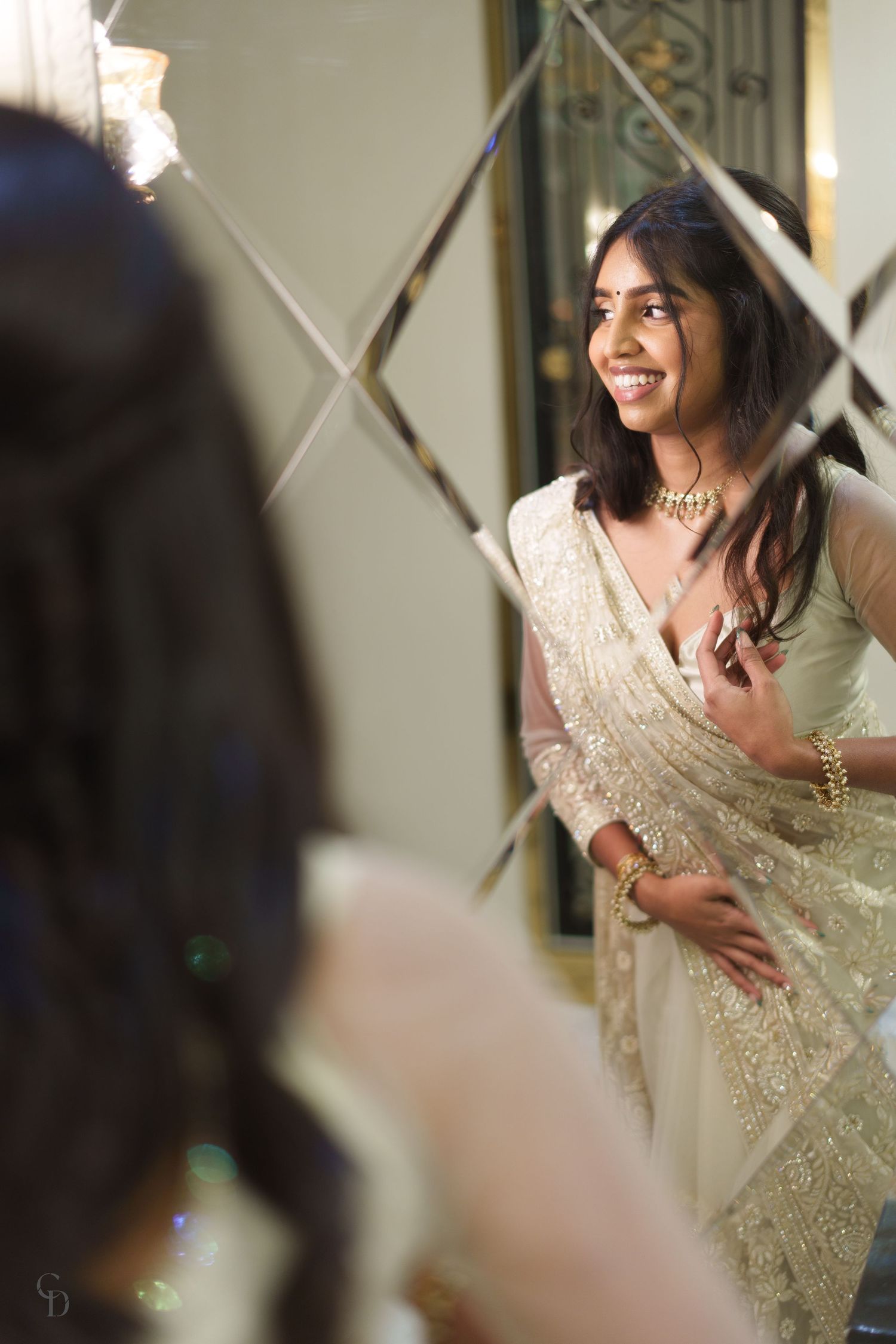 A traditional Indian bride smiles joyfully in a mirror reflection while wearing an elegant cream and gold sari.