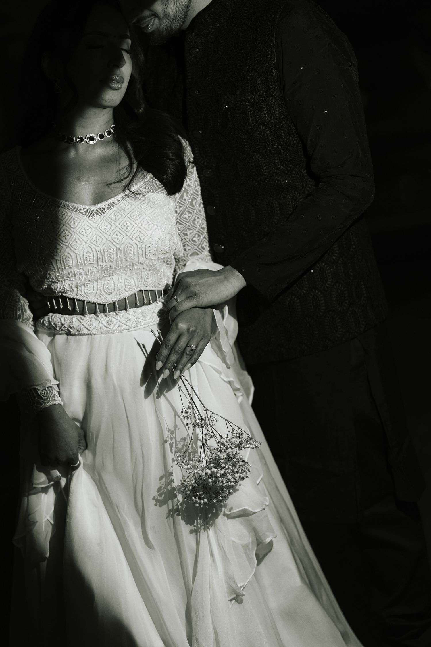 Black and white photo of a bride in white lace gown holding bouquet, embraced by groom in dark suit.