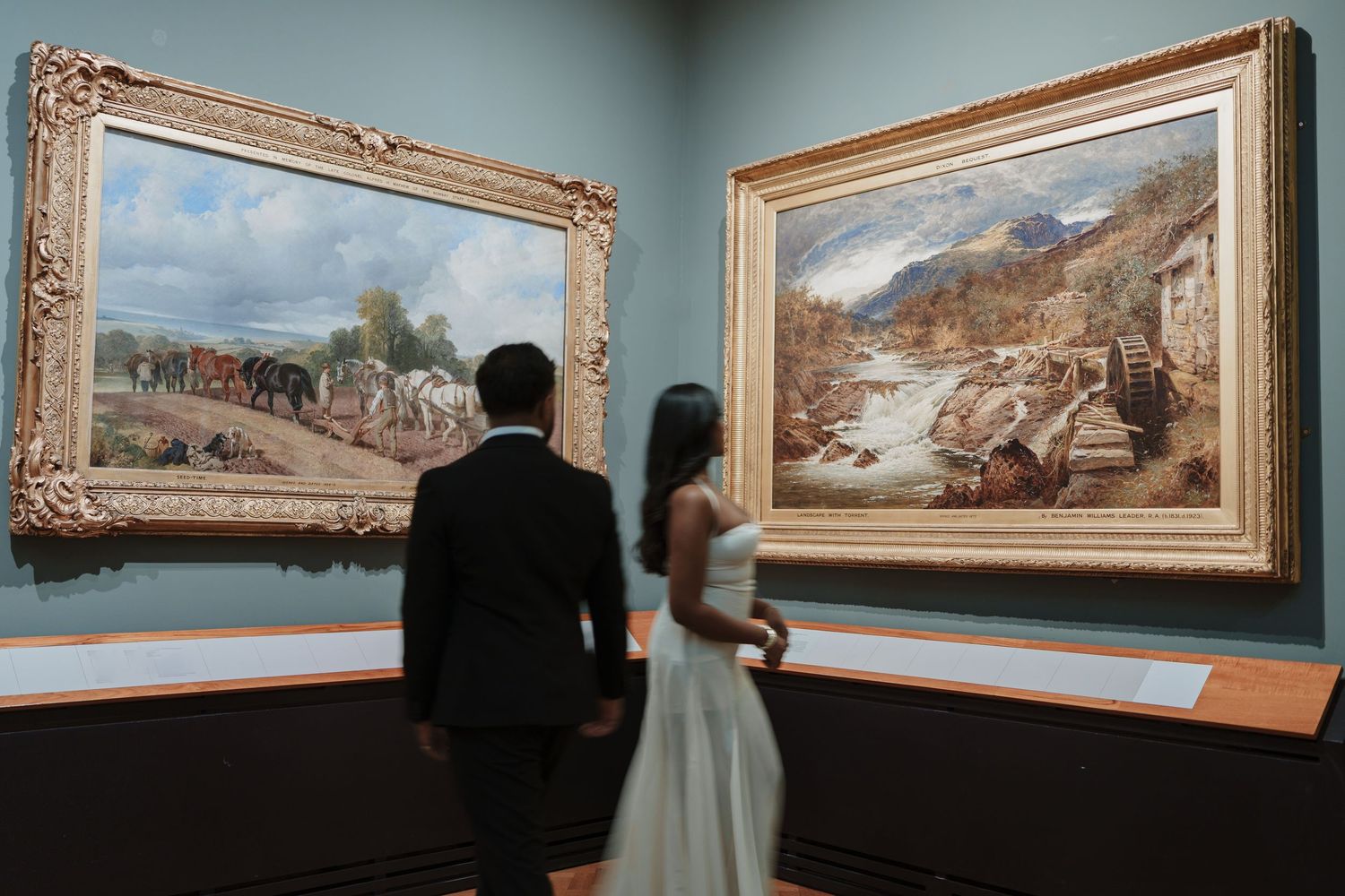 Couple in wedding attire viewing classic landscape paintings in an elegant art gallery museum.