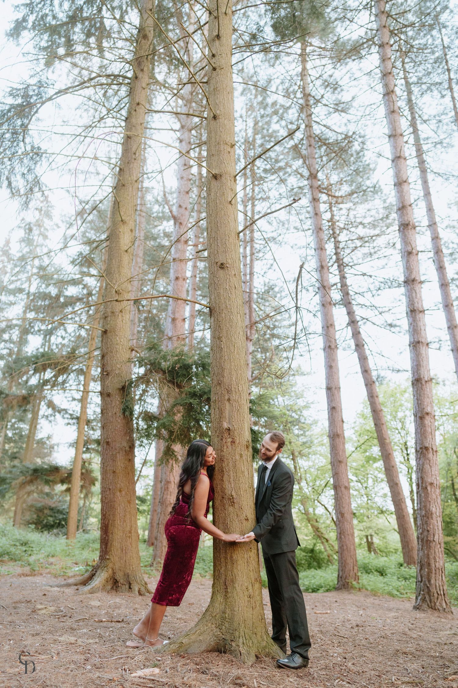 Playful engagement photo of couple posing among tall pine trees in forest.