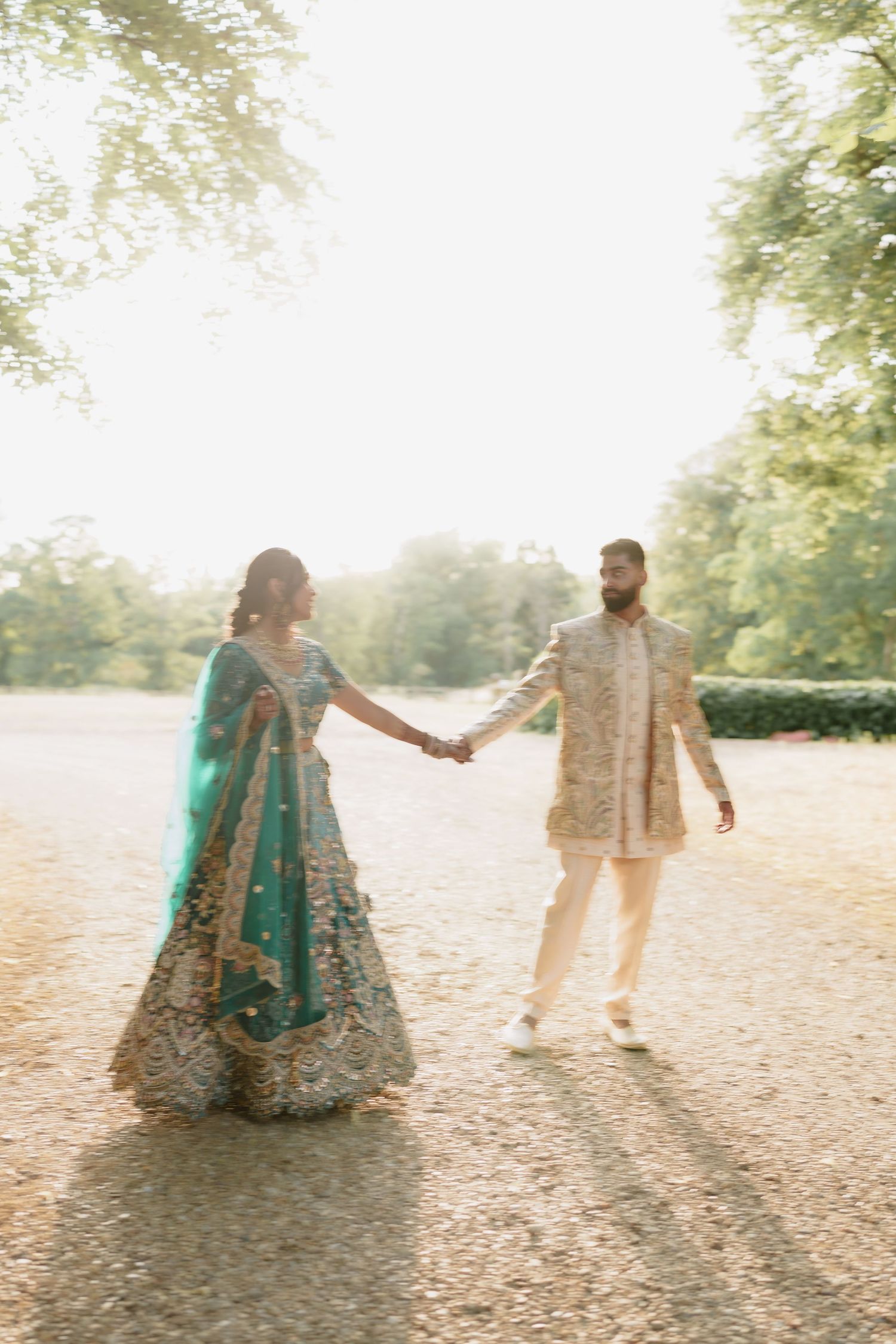 Couple in traditional Indian wedding attire walking through a sunlit park with haze creating a dreamy atmosphere.