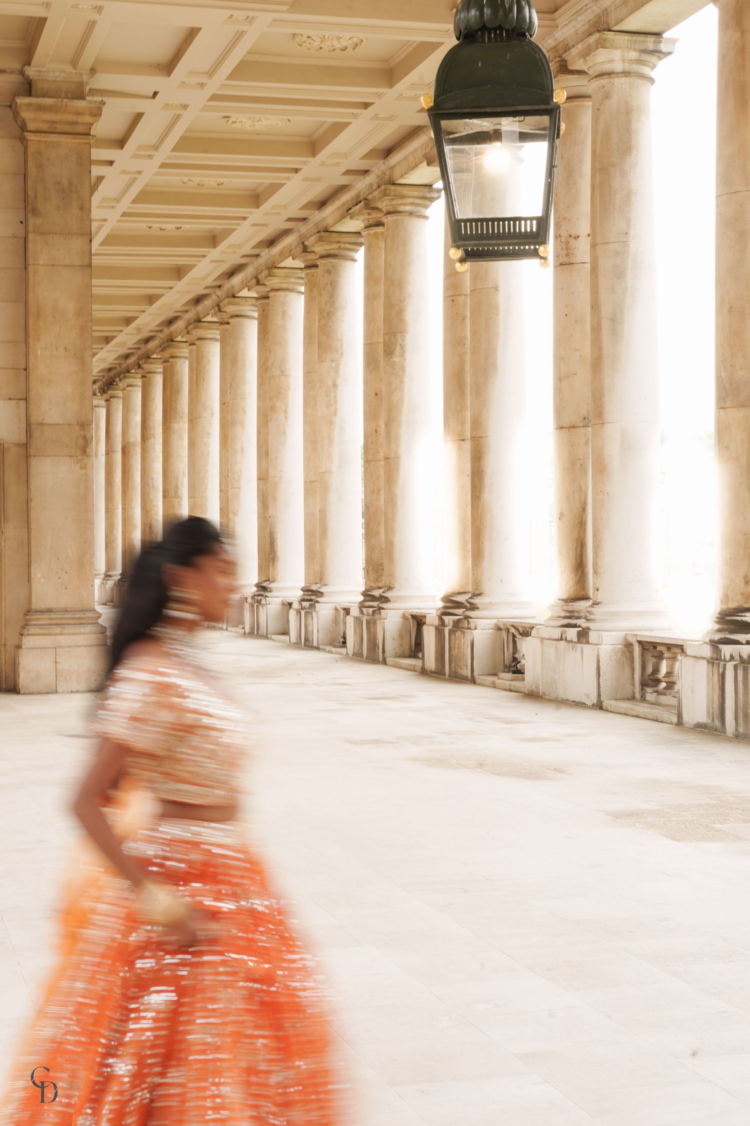 Motion blur photograph of someone in an orange dress walking through a colonnade with classical pillars.