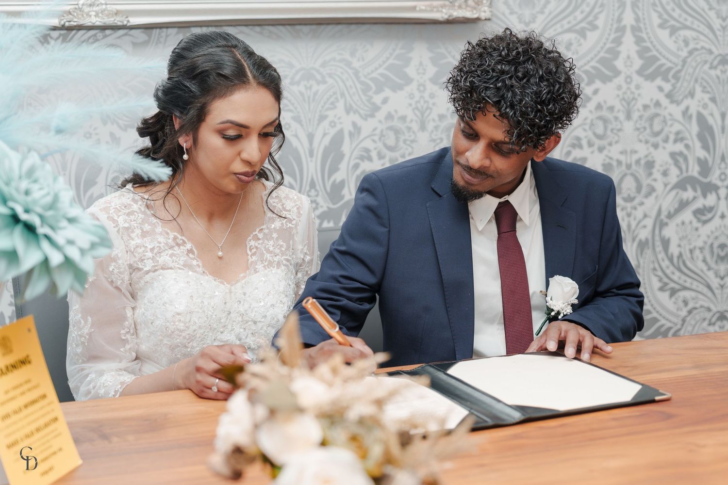 Wedding couple signing marriage documents at a table with decorative floral arrangements.