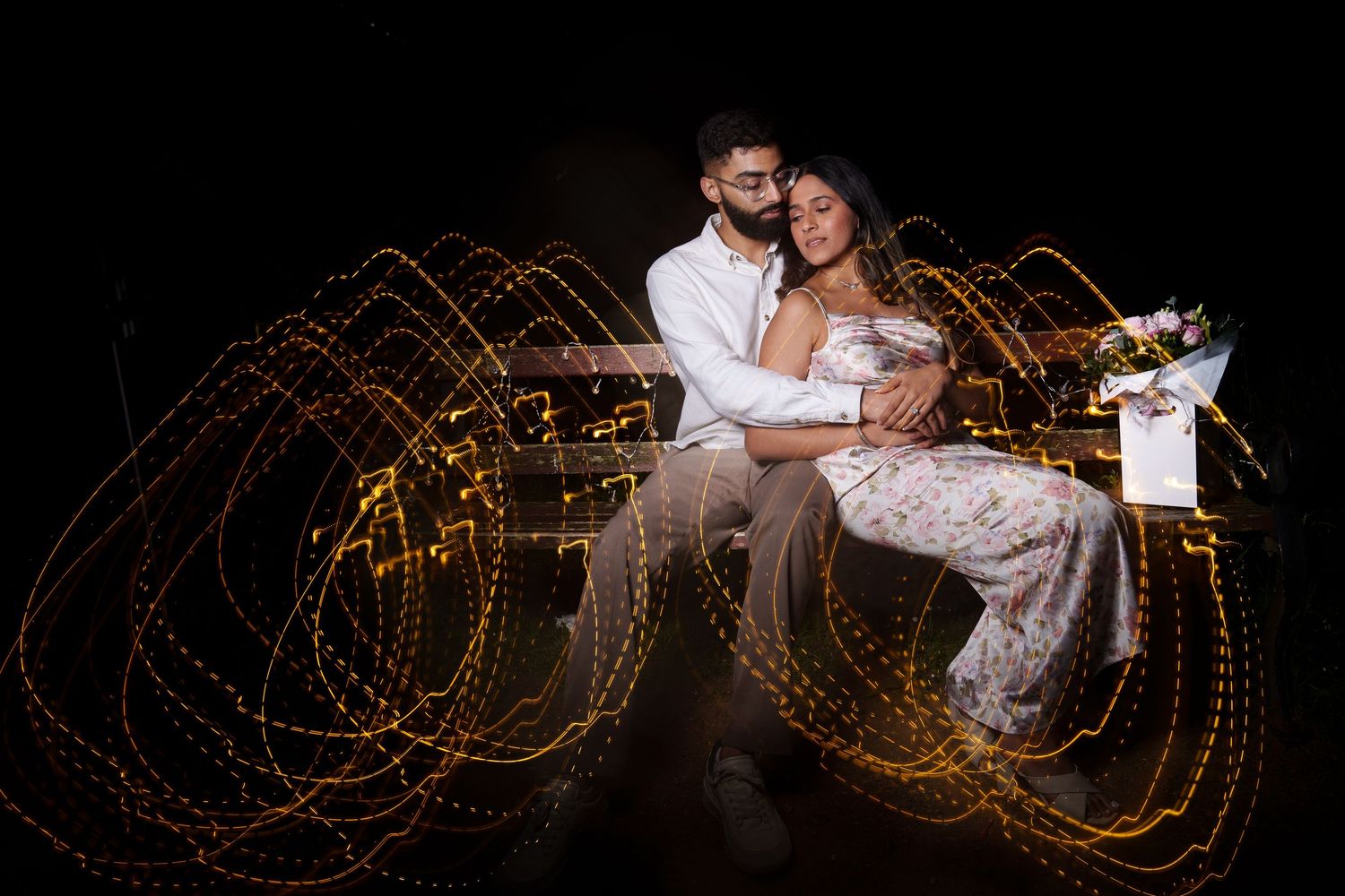 Couple poses against dark background with swirling light trails creating magical effect.
