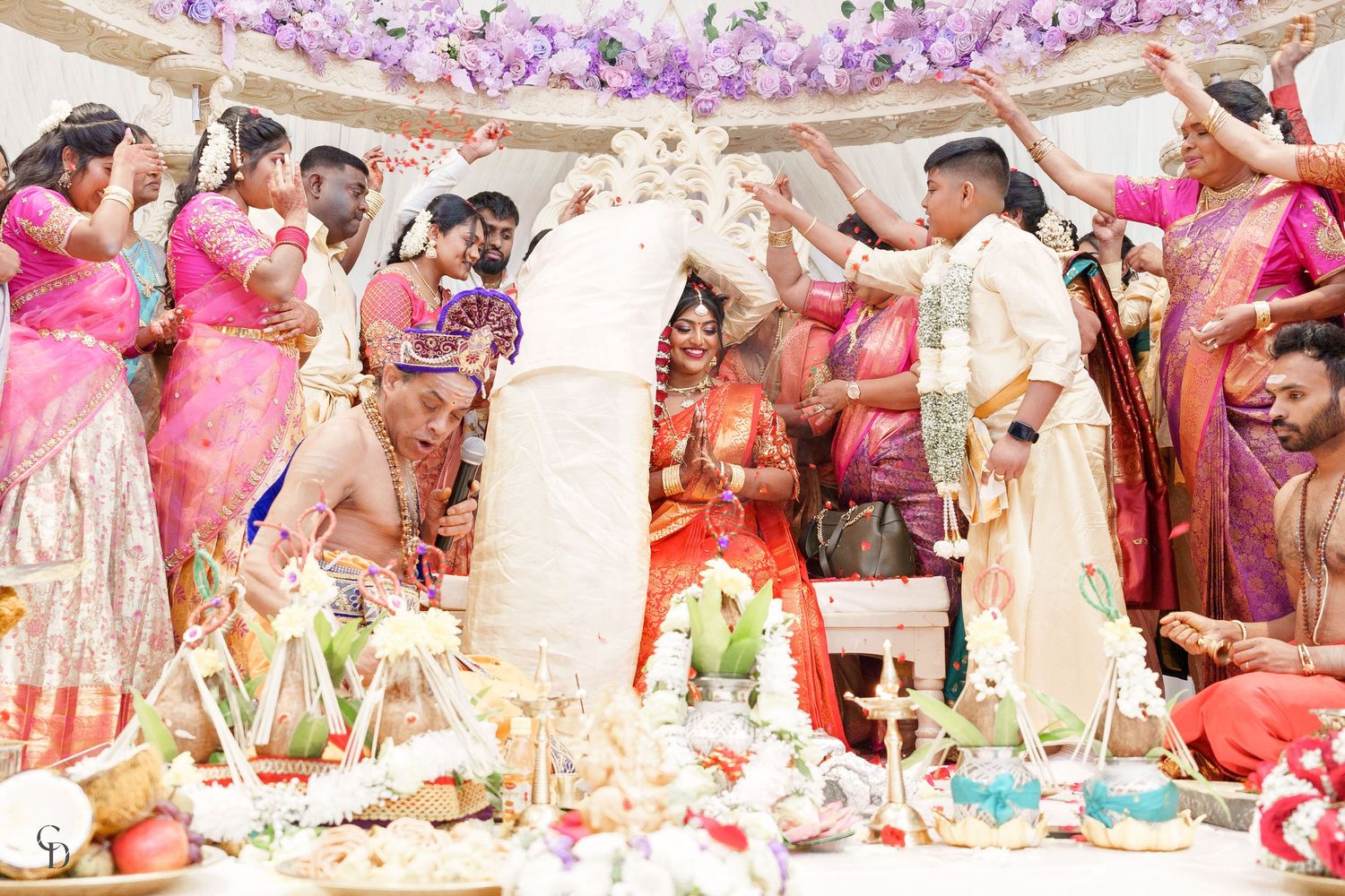 Traditional wedding ceremony with guests gathered around ritual items under purple floral decorations.