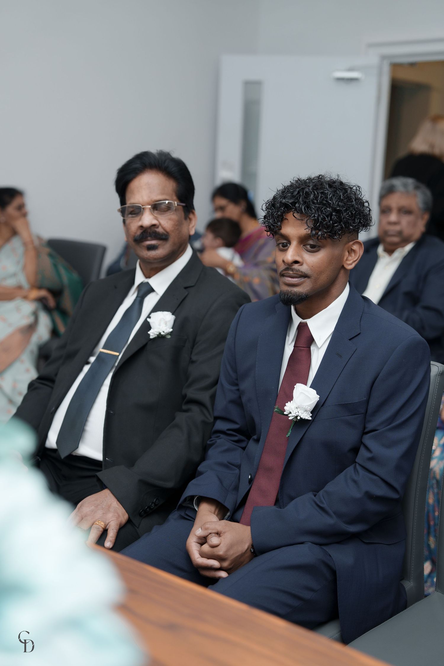 Two guests in formal attire sitting in a row during an indoor wedding ceremony.
