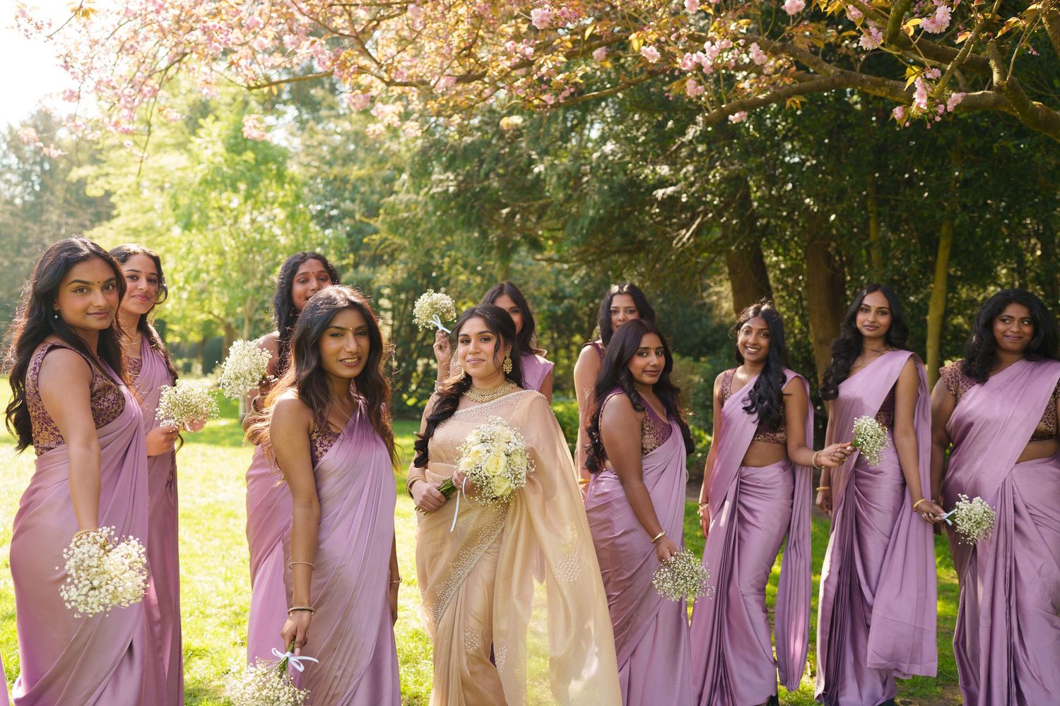 Bride in gold saree with bridesmaids in lilac dresses holding bouquets outdoors under blooming trees.