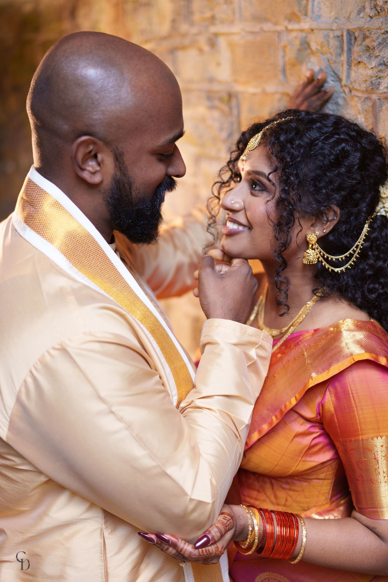 An intimate moment between newlyweds wearing traditional South Asian wedding attire against stone walls.