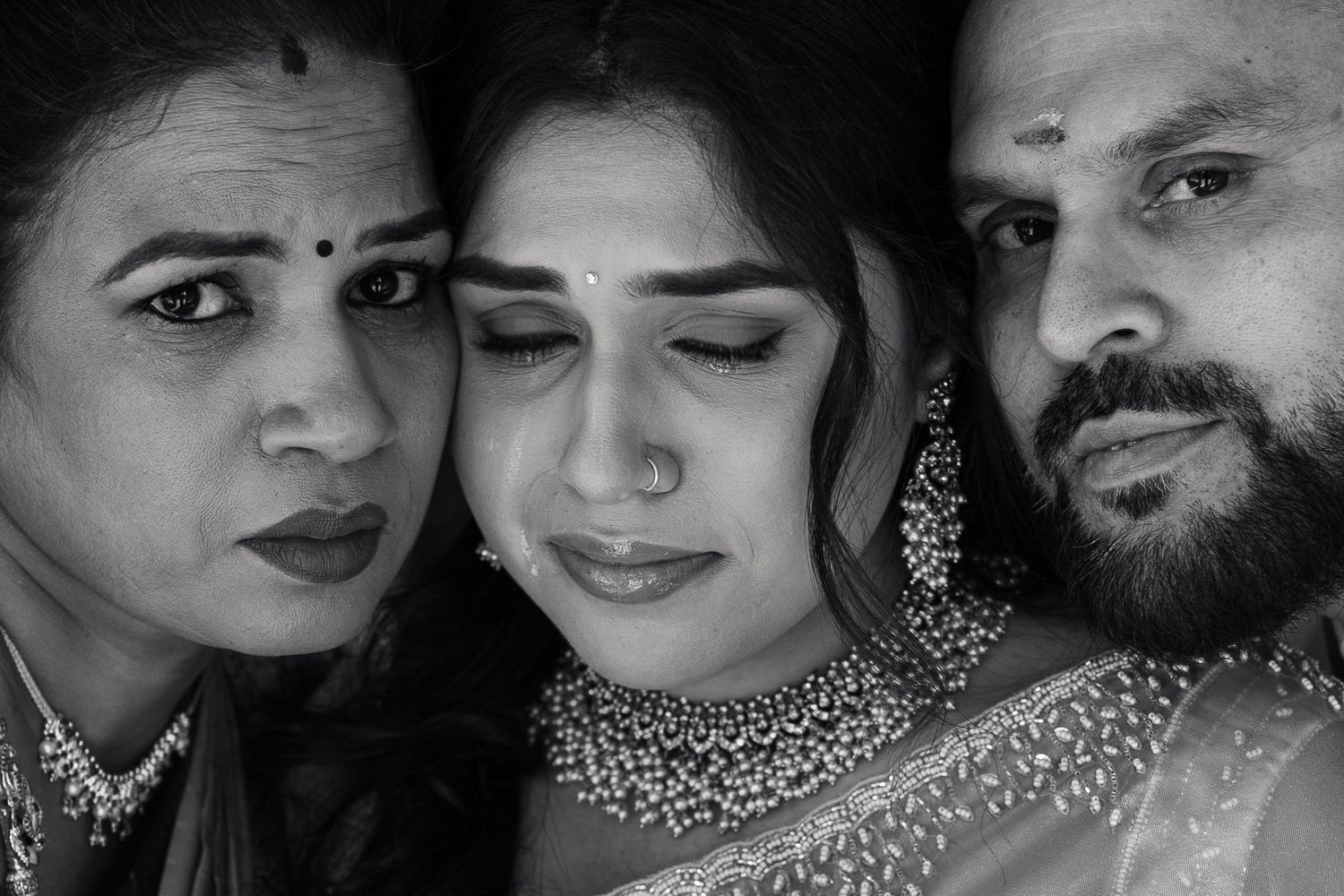 Black and white portrait of three people in traditional Indian attire, woman in center wearing ornate jewelry.