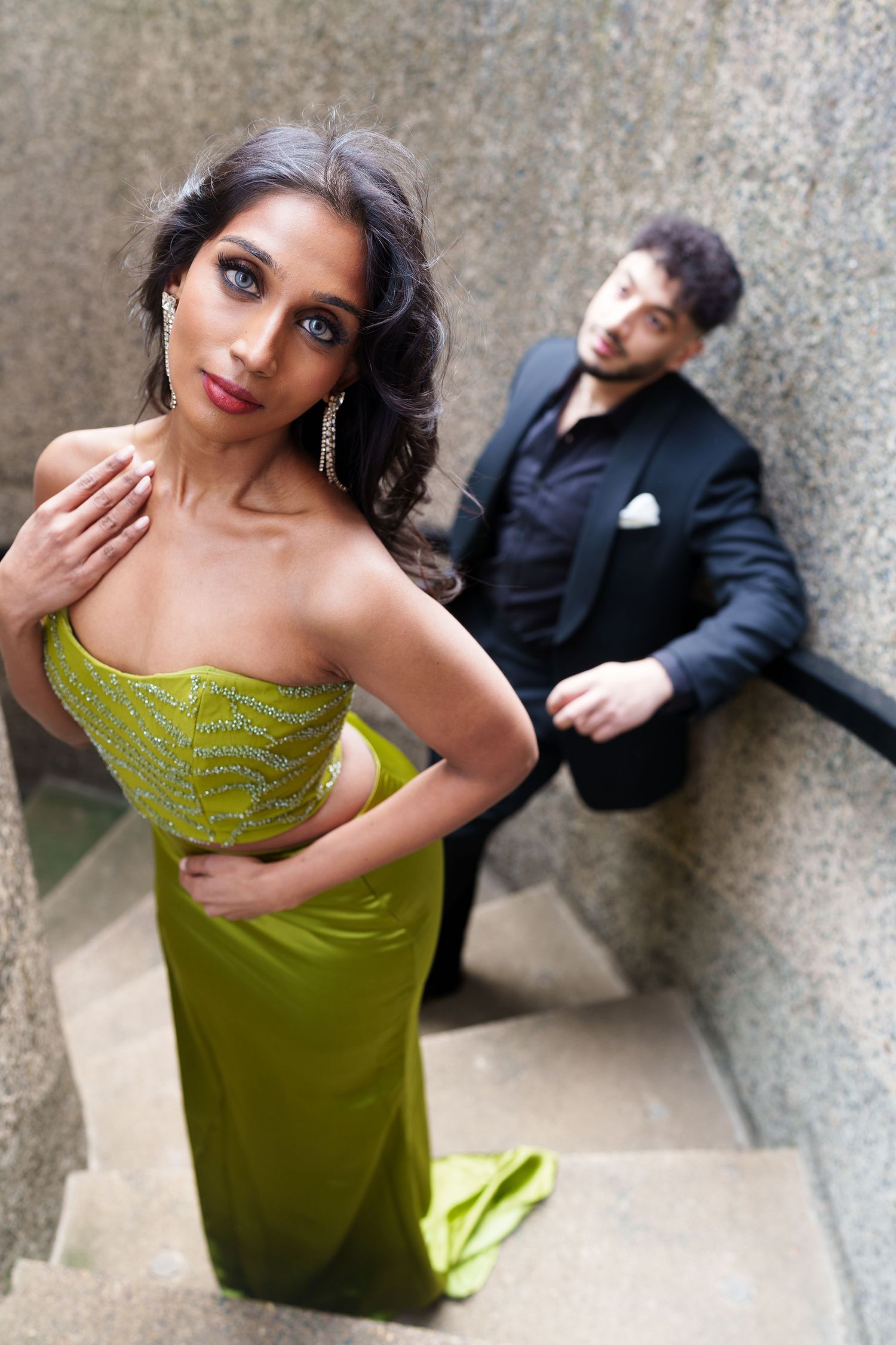 A glamorous couple poses on stone steps with the woman in a stunning green gown in the foreground.