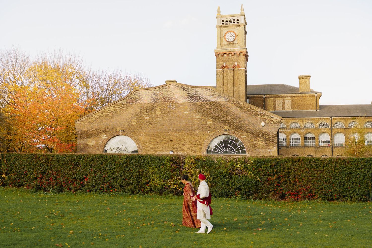 Couple in traditional attire posing on green lawn before historic brick building with clock tower in autumn.