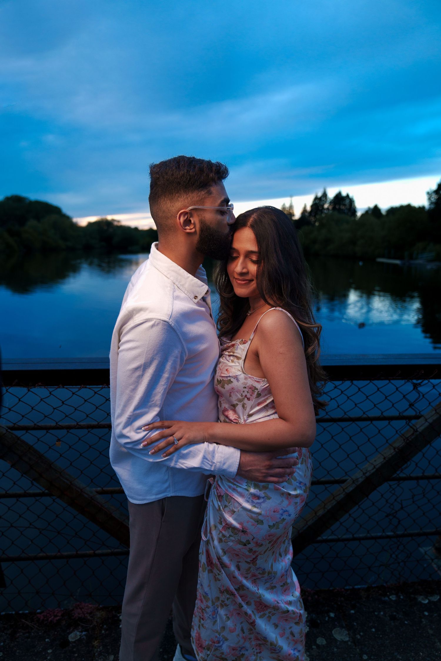 Couple embracing on bridge at twilight with serene lake backdrop.