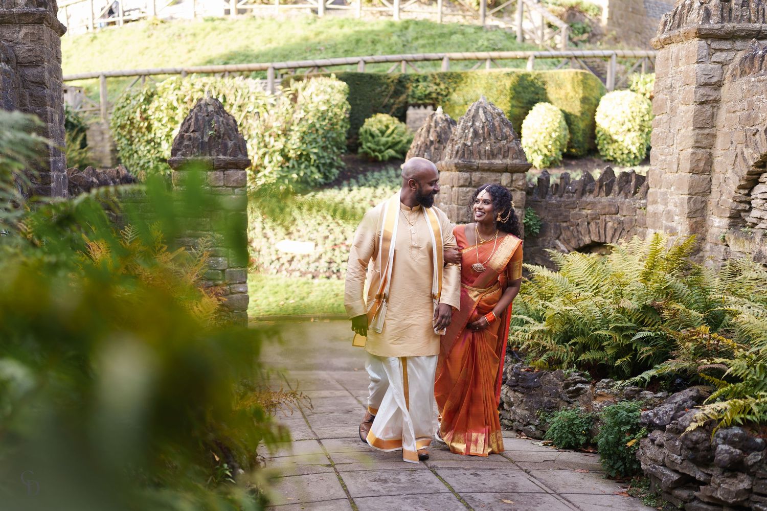 A couple walks hand in hand along a stone path surrounded by lush gardens and castle walls.
