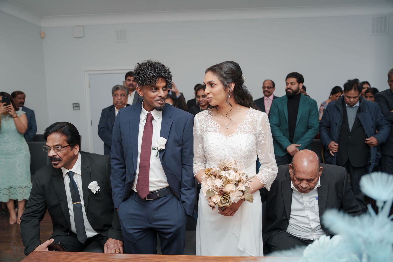 Wedding ceremony guests and couple gathered in a bright indoor venue with white walls.