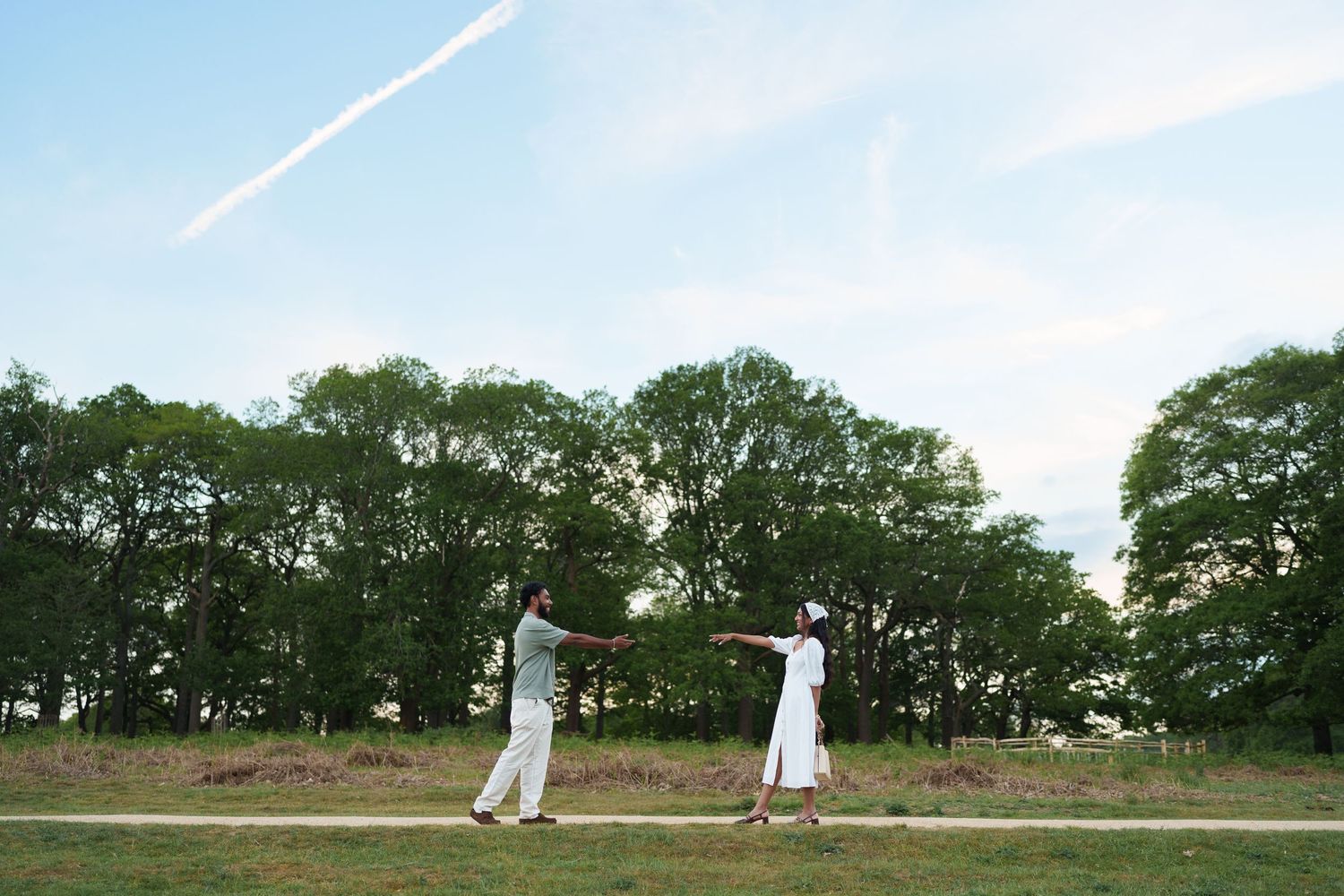Couple in white outfits dancing outdoors on a path surrounded by lush green trees under a blue sky.