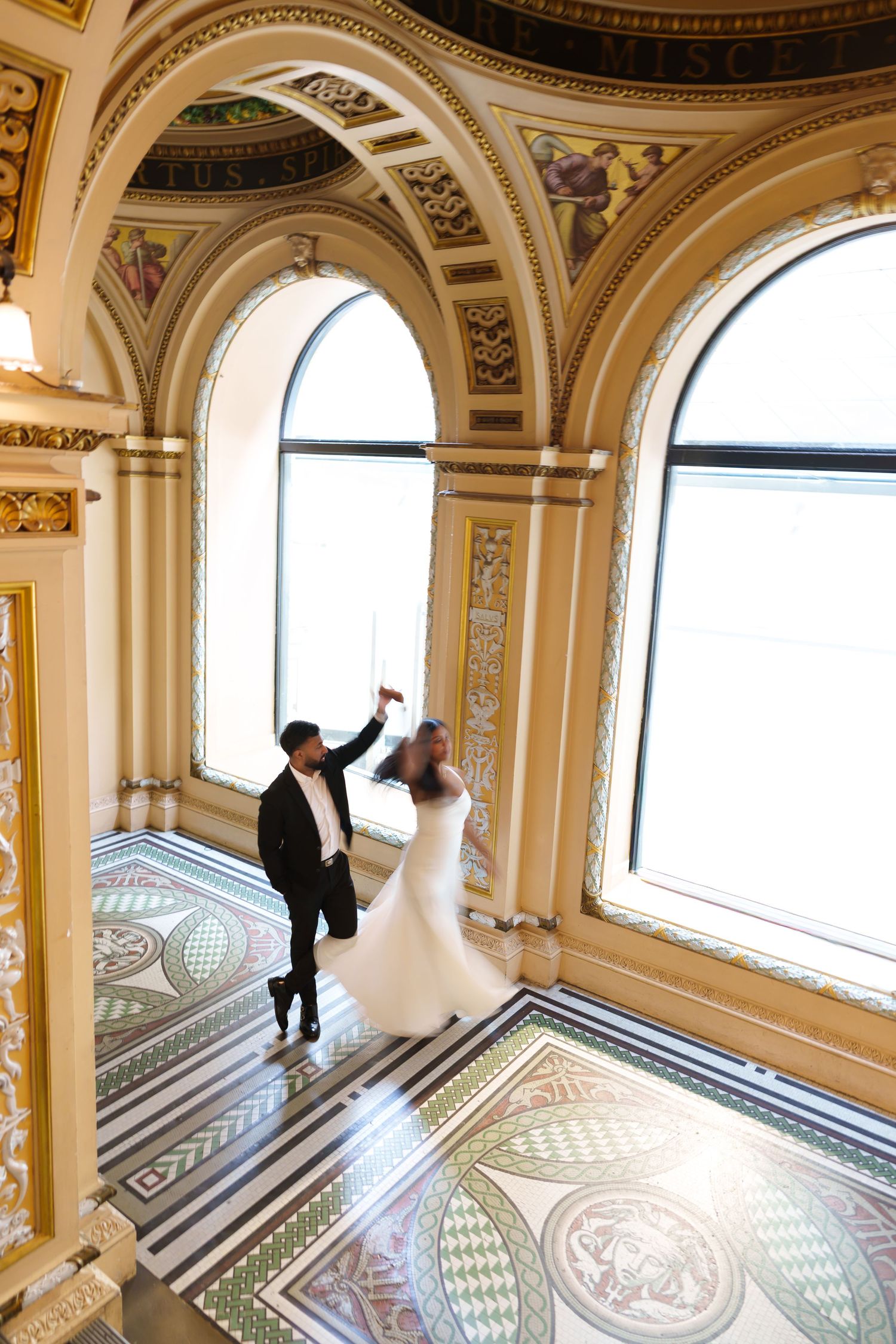 Bride and groom dancing in an ornate hall with arched windows and decorative mosaic tile floors.