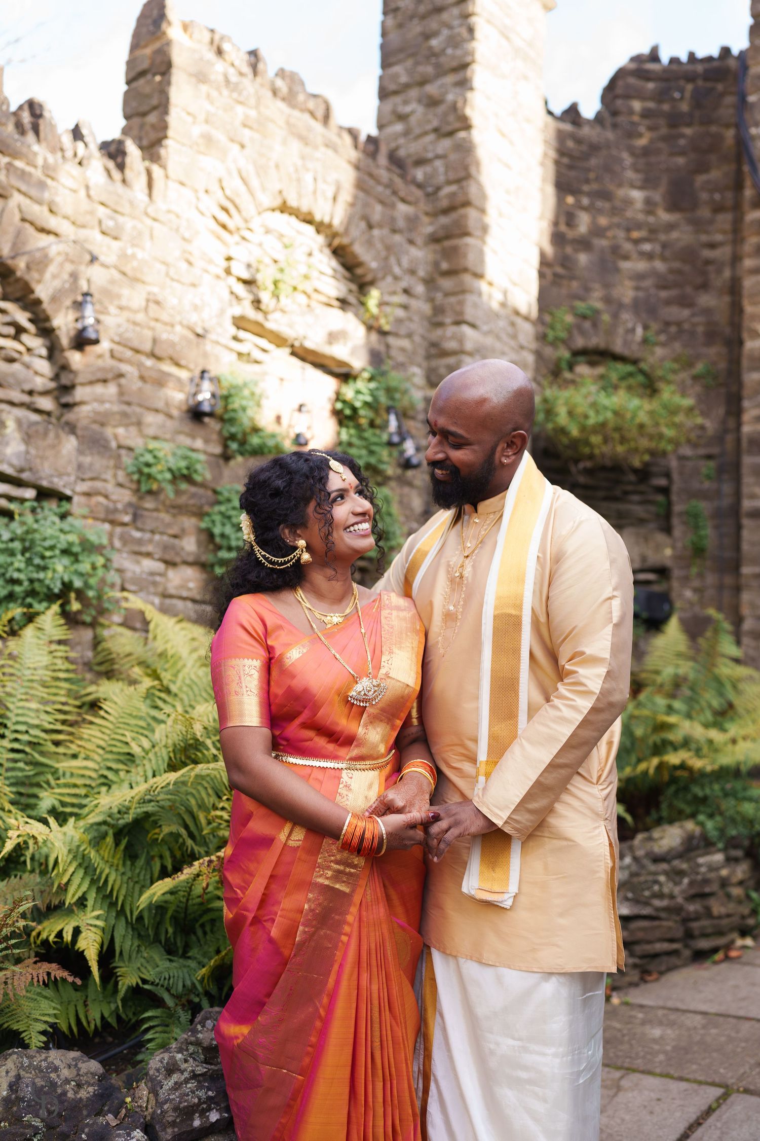 A couple in traditional South Asian wedding attire standing together against stone castle ruins.